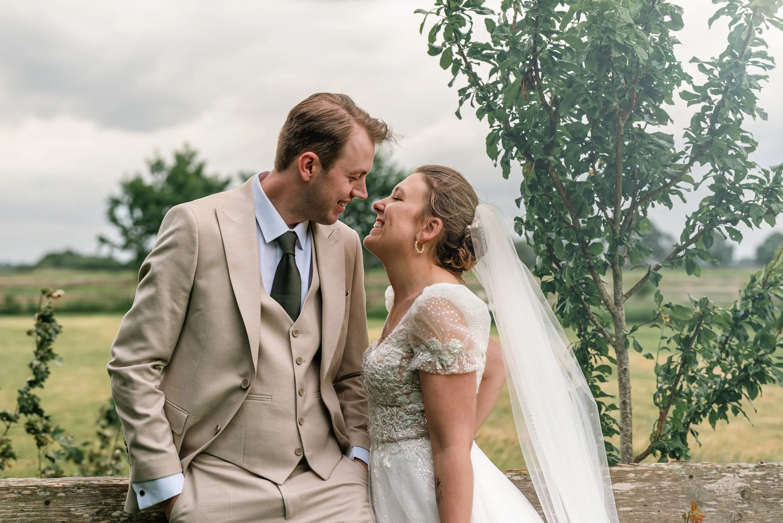 A bride and groom in wedding attire sharing a joyful moment outdoors on a cloudy day, with trees and greenery in the background.