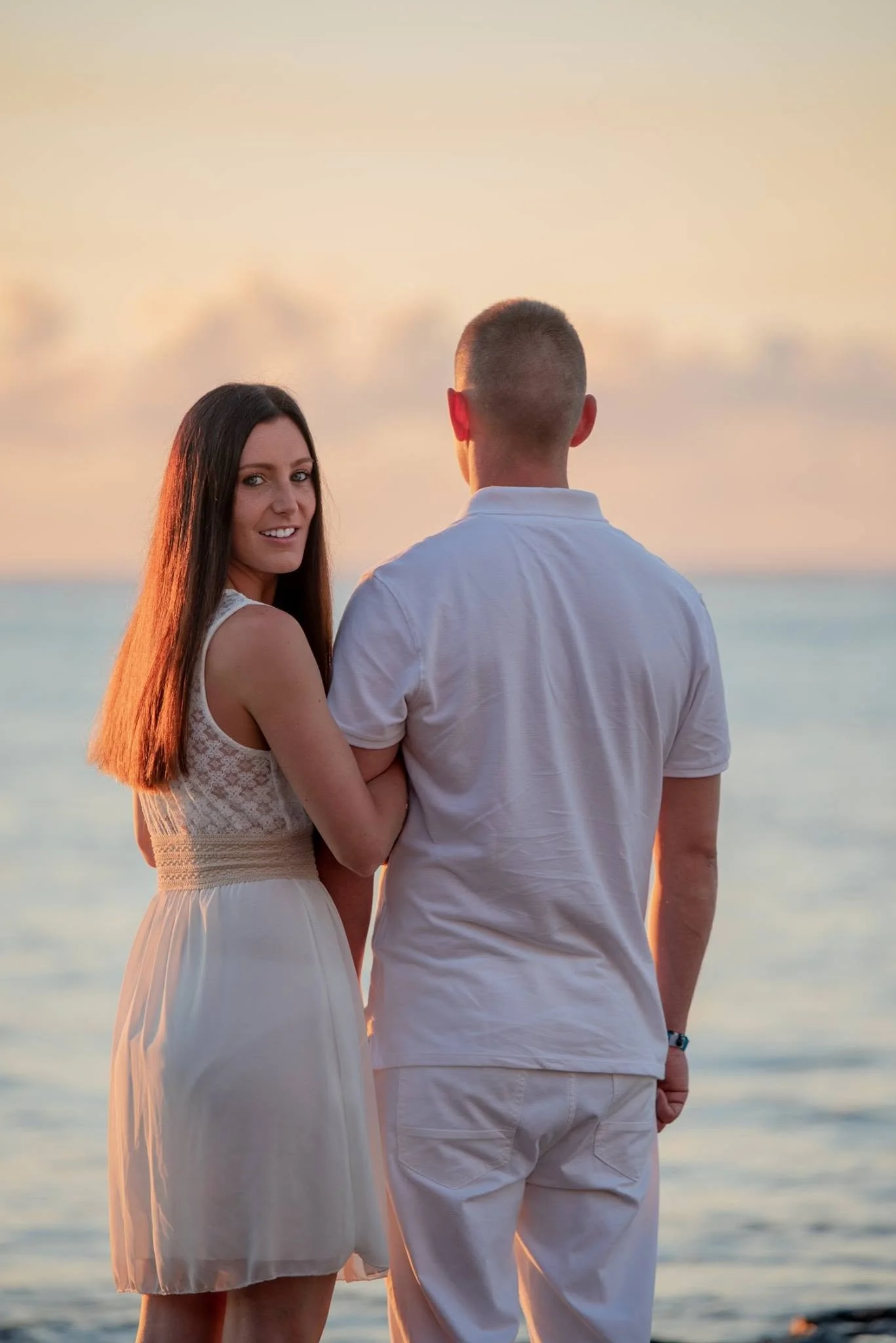 A woman and a man facing the ocean at sunset, with the woman smiling at the camera and the man with his back turned, standing close together.