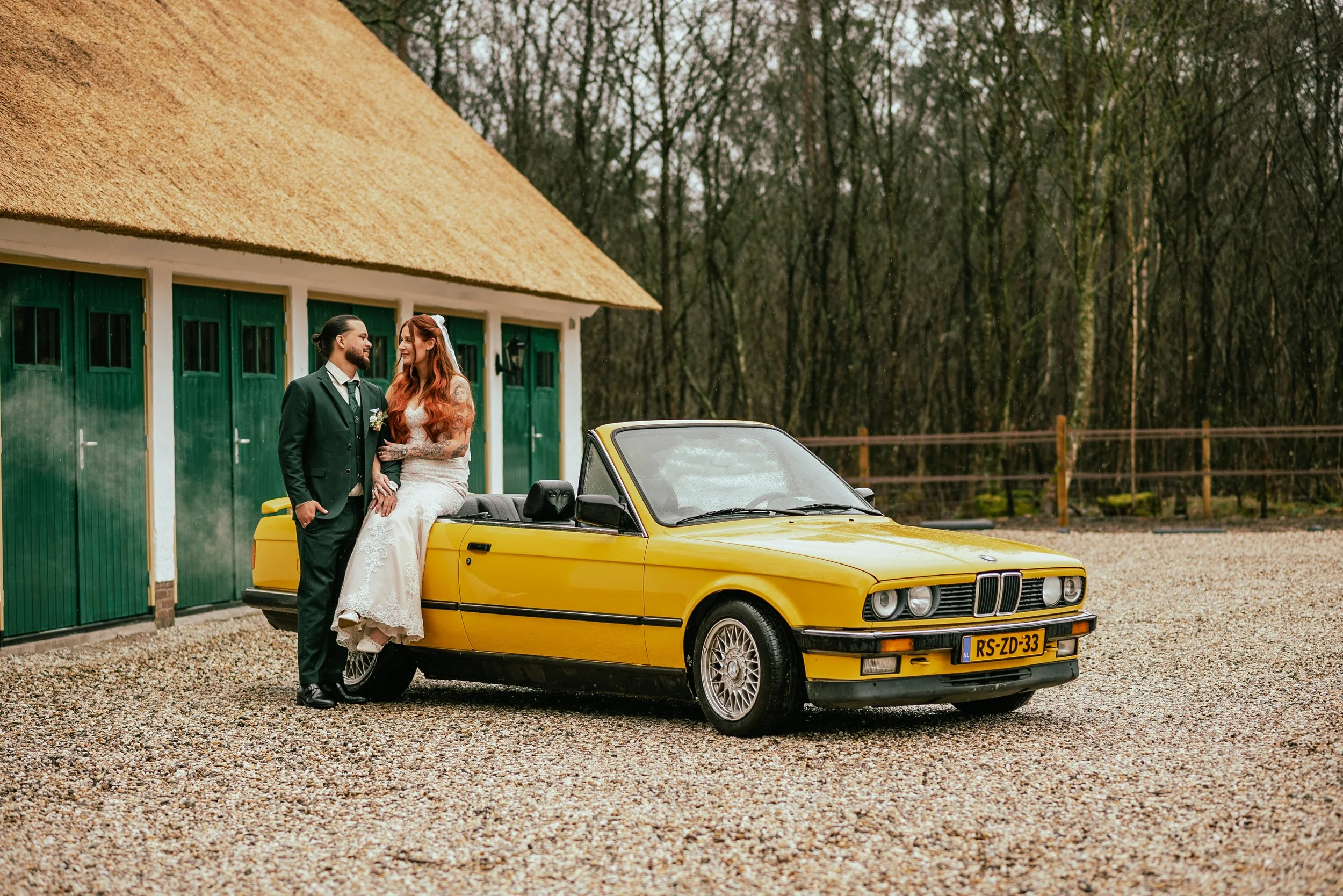 A bride and groom standing next to a yellow vintage BMW car outside a green barn with a thatched roof, with trees in the background.