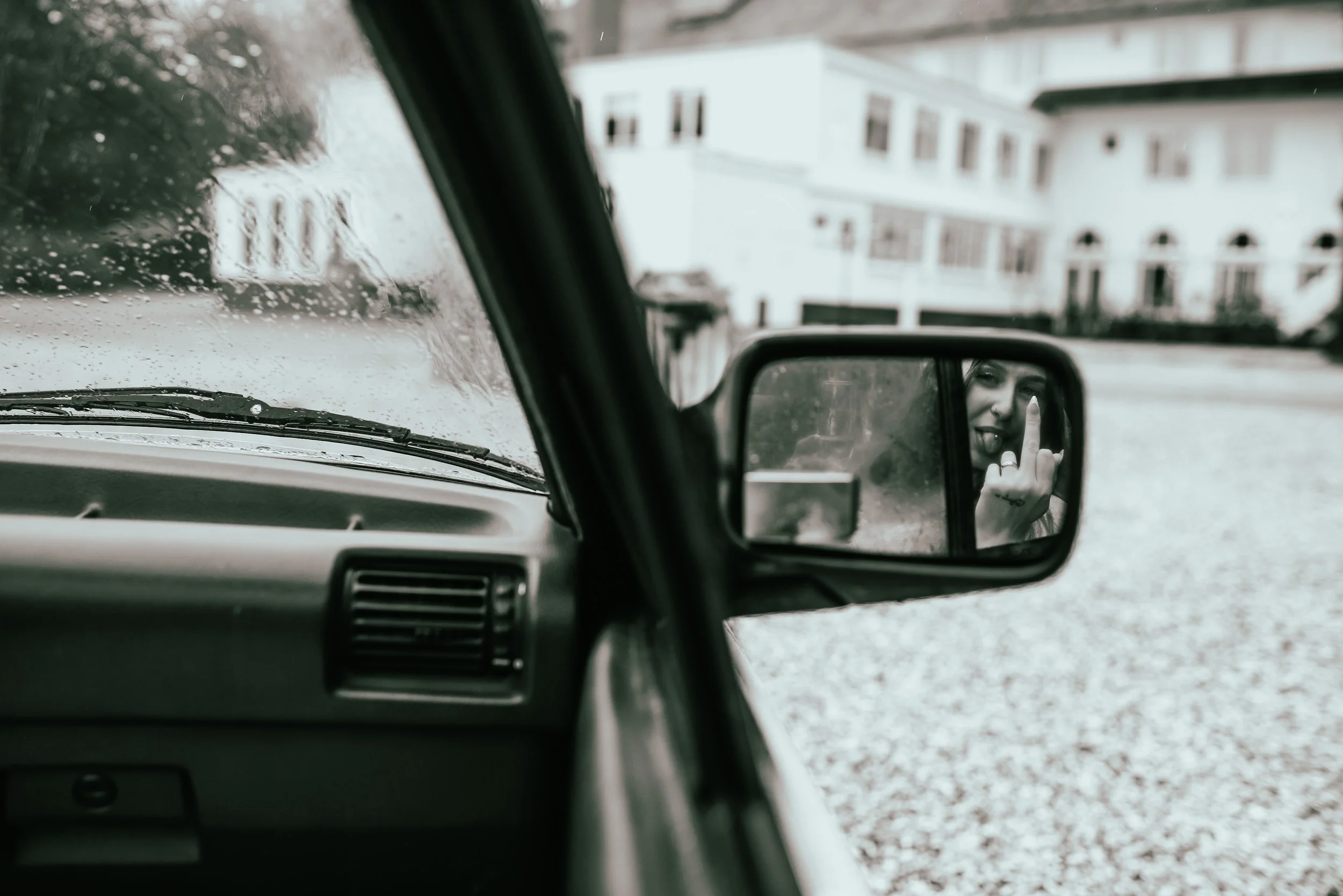 A woman is taking a selfie in the side mirror of a car during a rainy day, with raindrops visible on the window and a white house in the background.