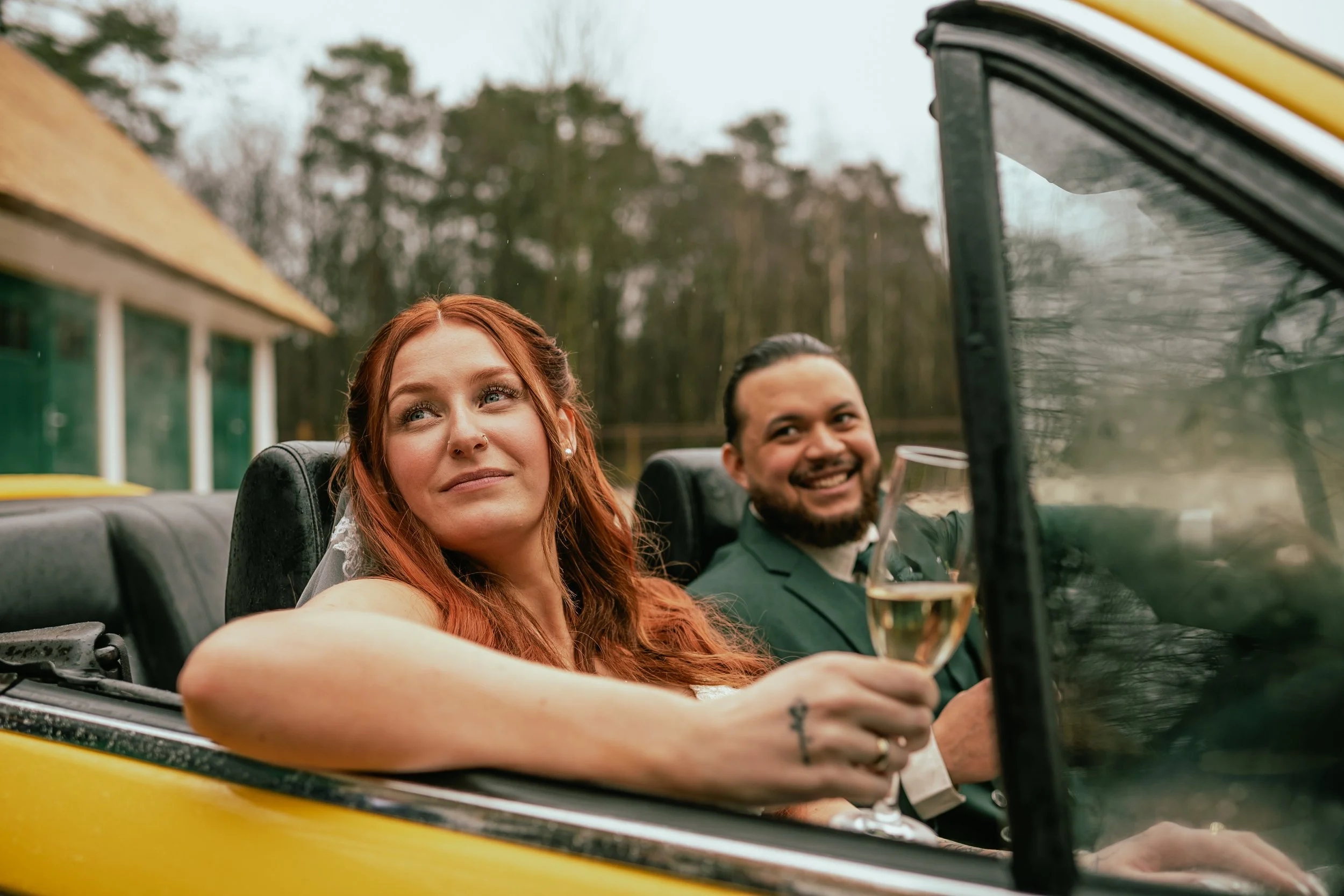 A smiling woman with red hair and a man with dark hair in a green suit, both enjoying a ride in a convertible car, holding drinks, with trees and a building in the background.