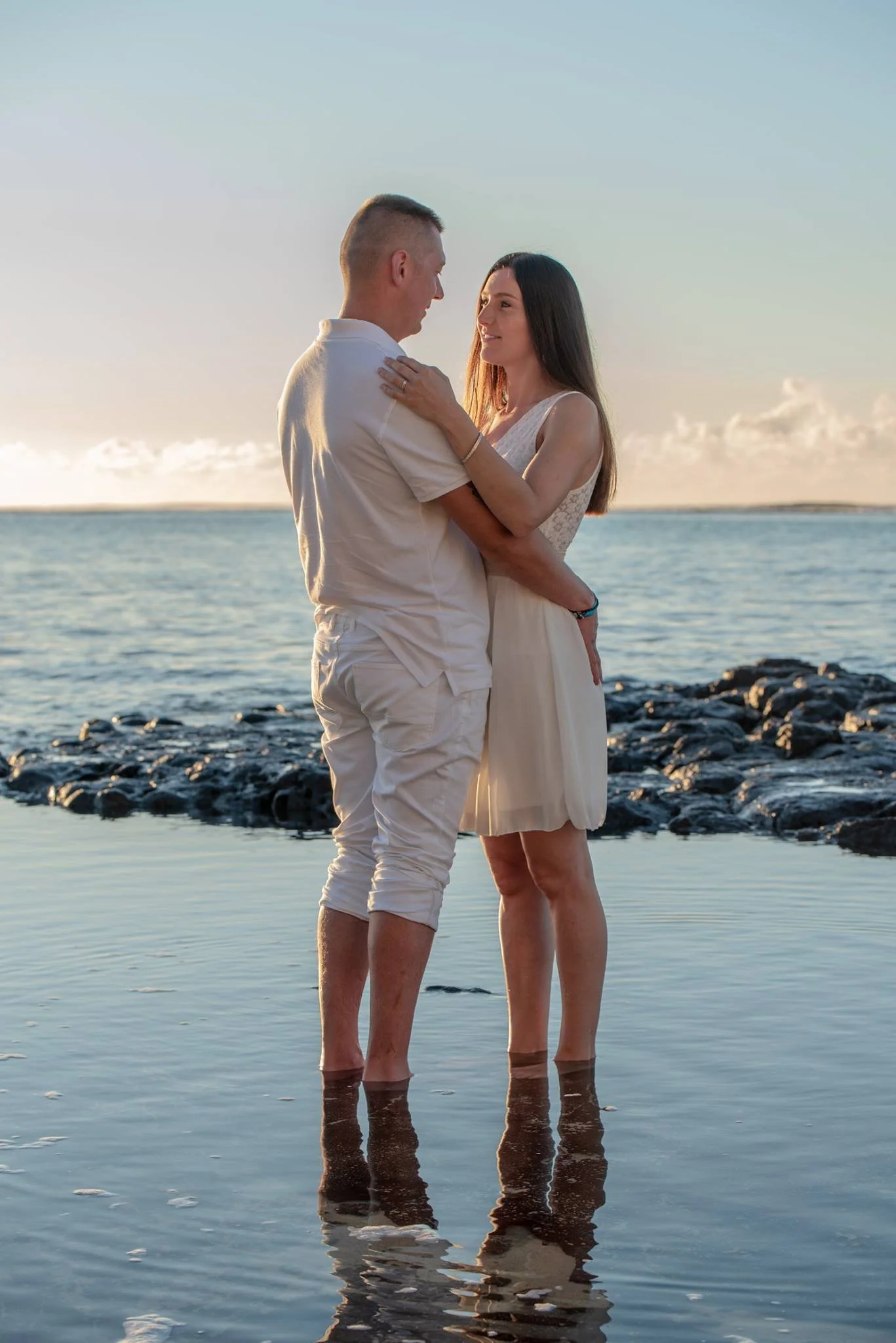 A couple stands in shallow ocean water during sunset, facing each other lovingly with their arms around each other.