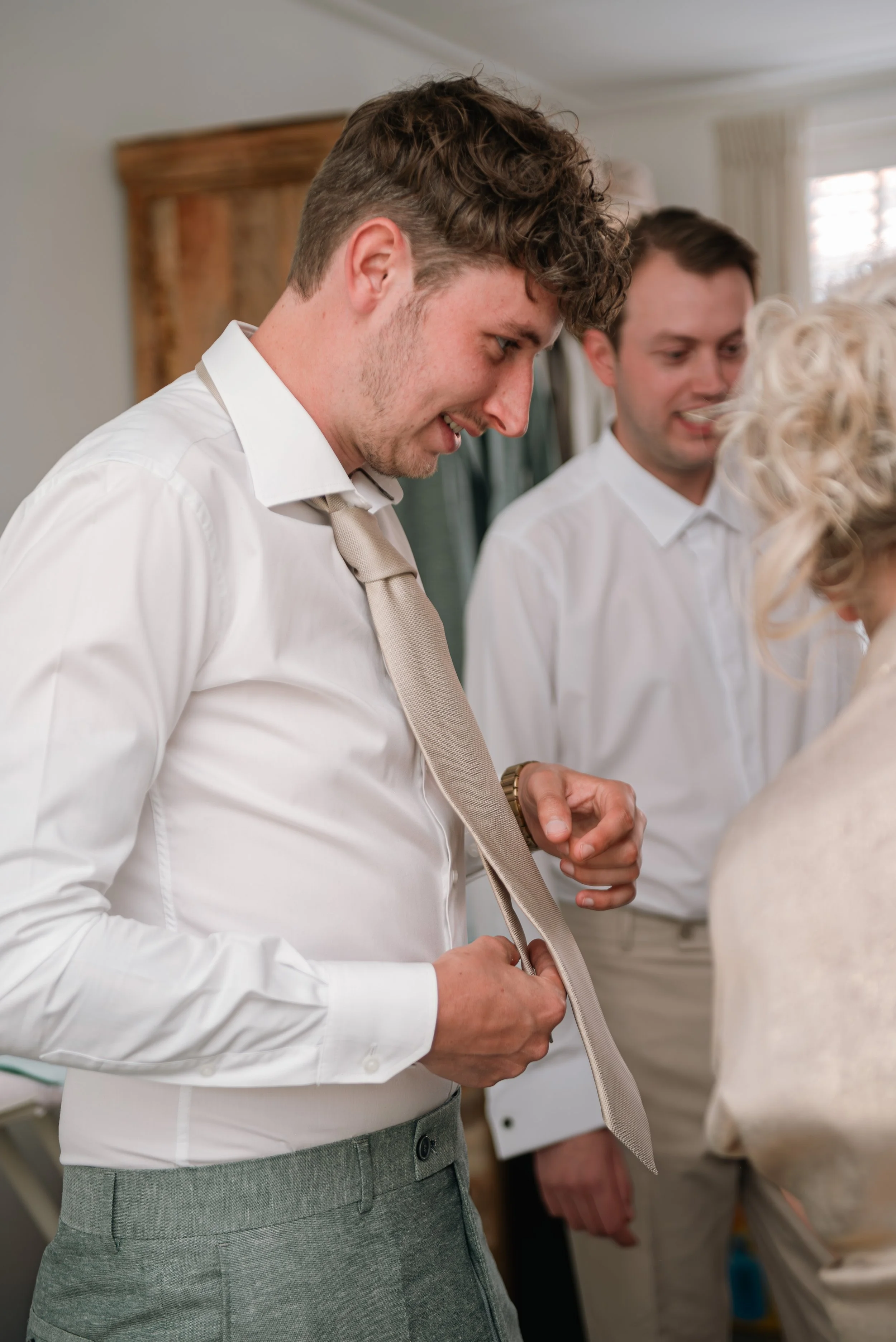 A man with brown curly hair, wearing a white shirt and beige tie, is adjusting a beige tie, standing among others in an indoor setting.