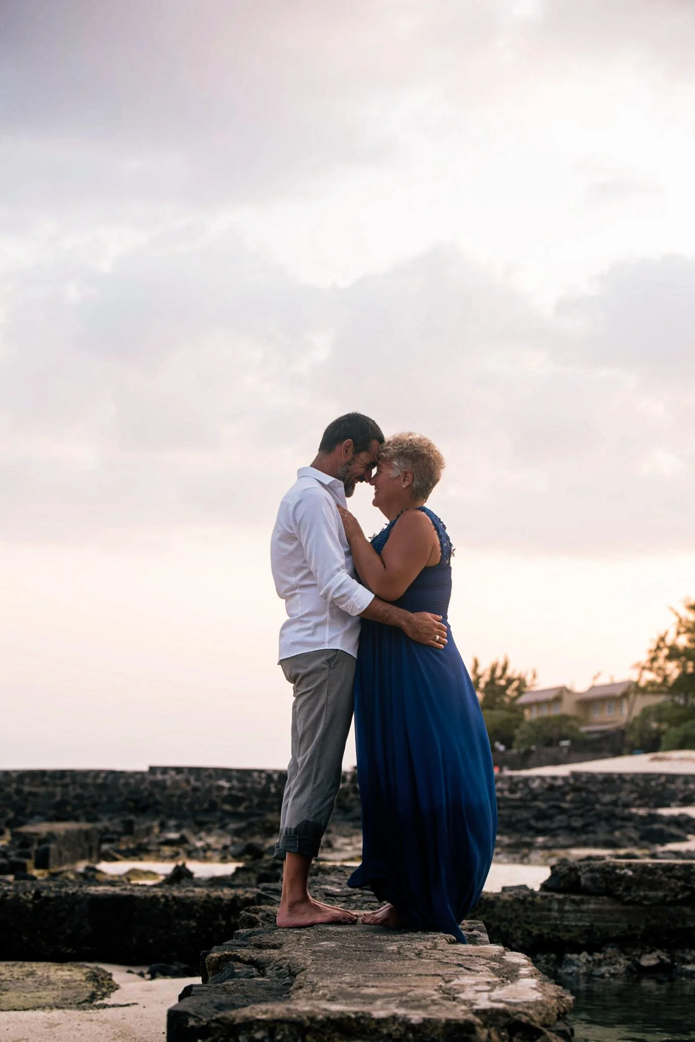 A couple stands close together on a rocky shore, foreheads touching and eyes closed, embracing each other during sunset or twilight, with houses and trees in the background.