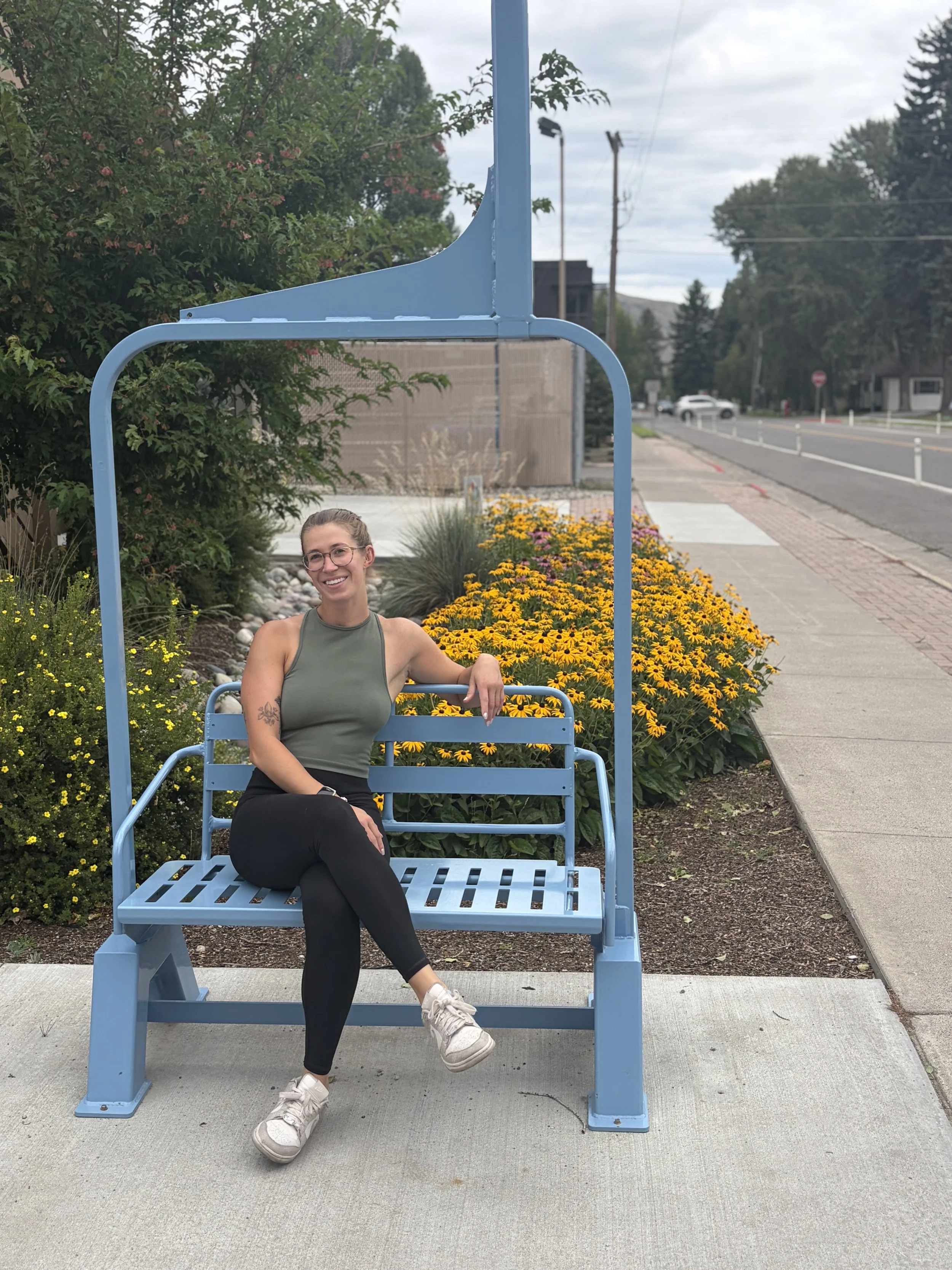 A woman with glasses and a tattoo on her right arm sitting on a blue metal bench with a large backrest, smiling, outdoors near a flower bed with yellow and pink flowers, and a sidewalk.