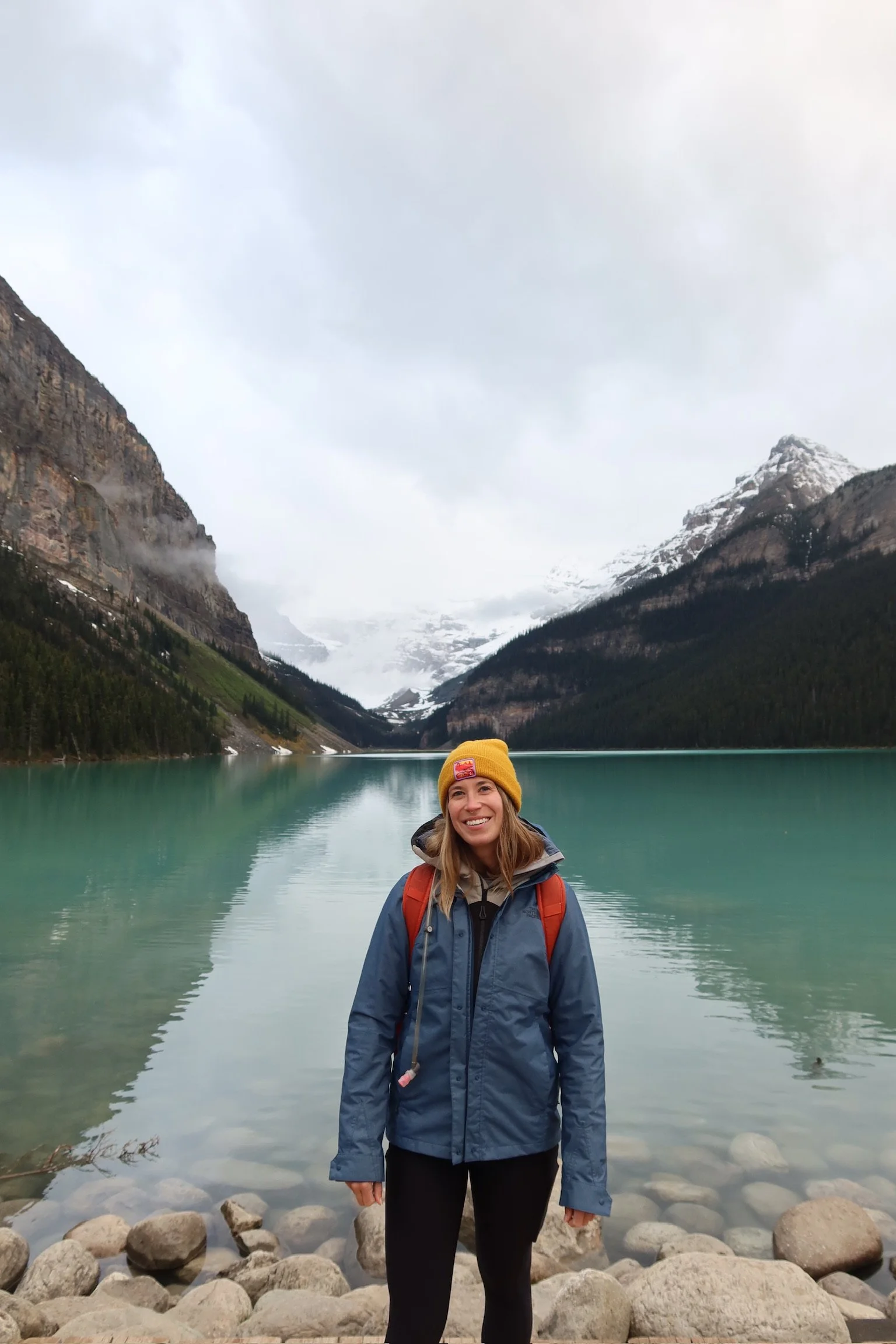 A smiling woman wearing a yellow beanie, blue jacket, and black pants standing on rocks at the edge of a serene mountain lake with snow-capped peaks in the background.