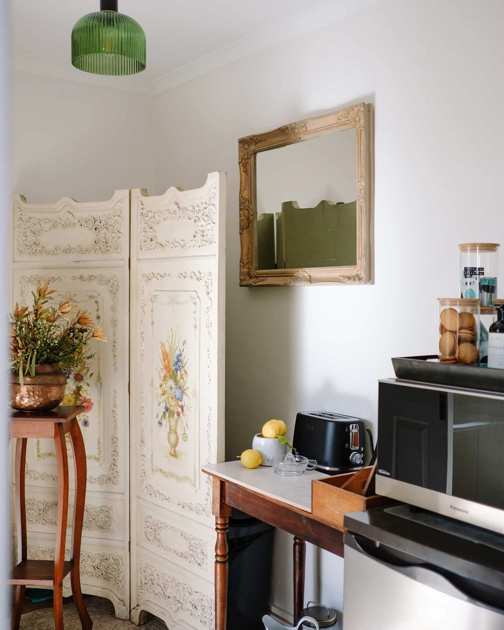 Interior of a cozy kitchen corner with a white ornate folding screen, a plant in a copper pot, a small wooden table, and a white wall. There is a large mirror with a decorative gold frame on the wall, and various kitchen appliances and utensils on a 