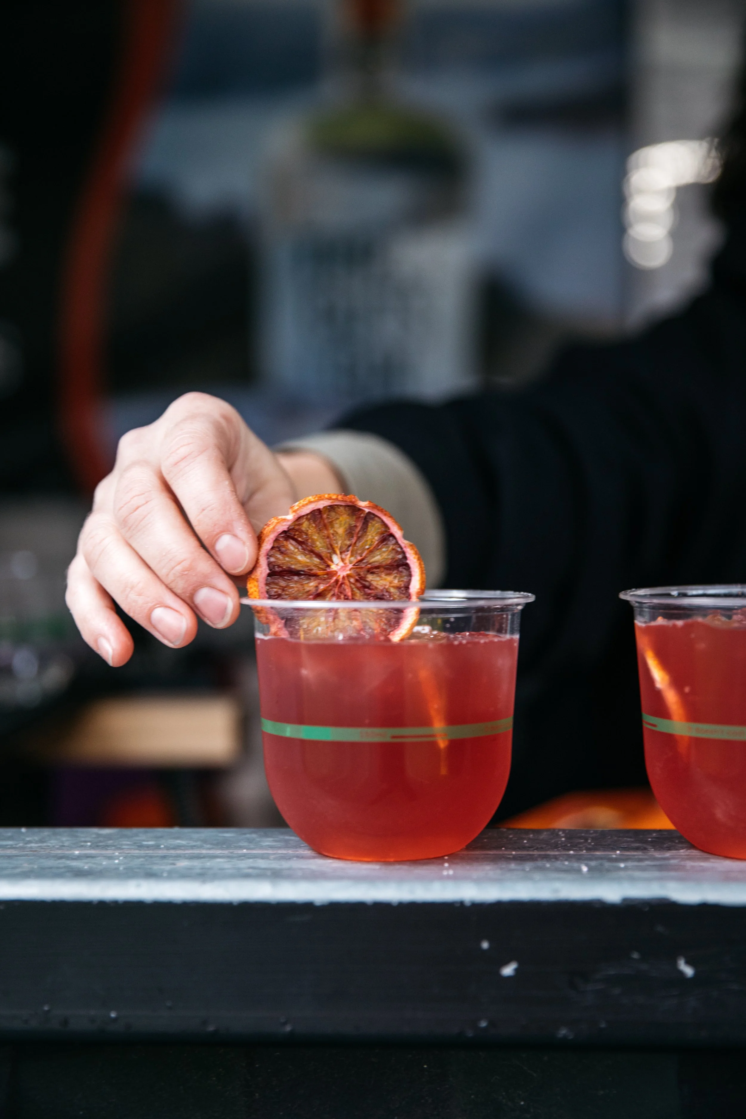 A person is holding a slice of dried blood orange above a glass of red-colored beverage with a slice of blood orange garnishing the rim.