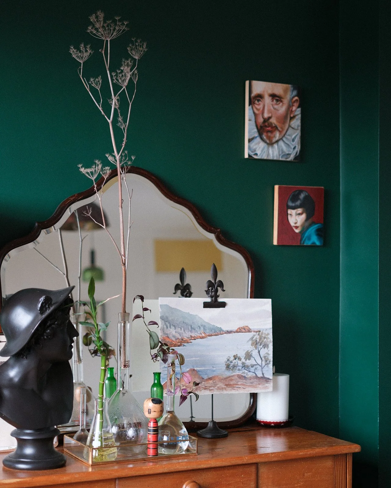 A vintage wooden dresser with a decorative mirror, featuring art supplies, glass bottles, a black bust sculpture, a painted landscape, and greenery inside a glass container. Two framed portraits hang on a dark green wall behind, and a paper lantern w
