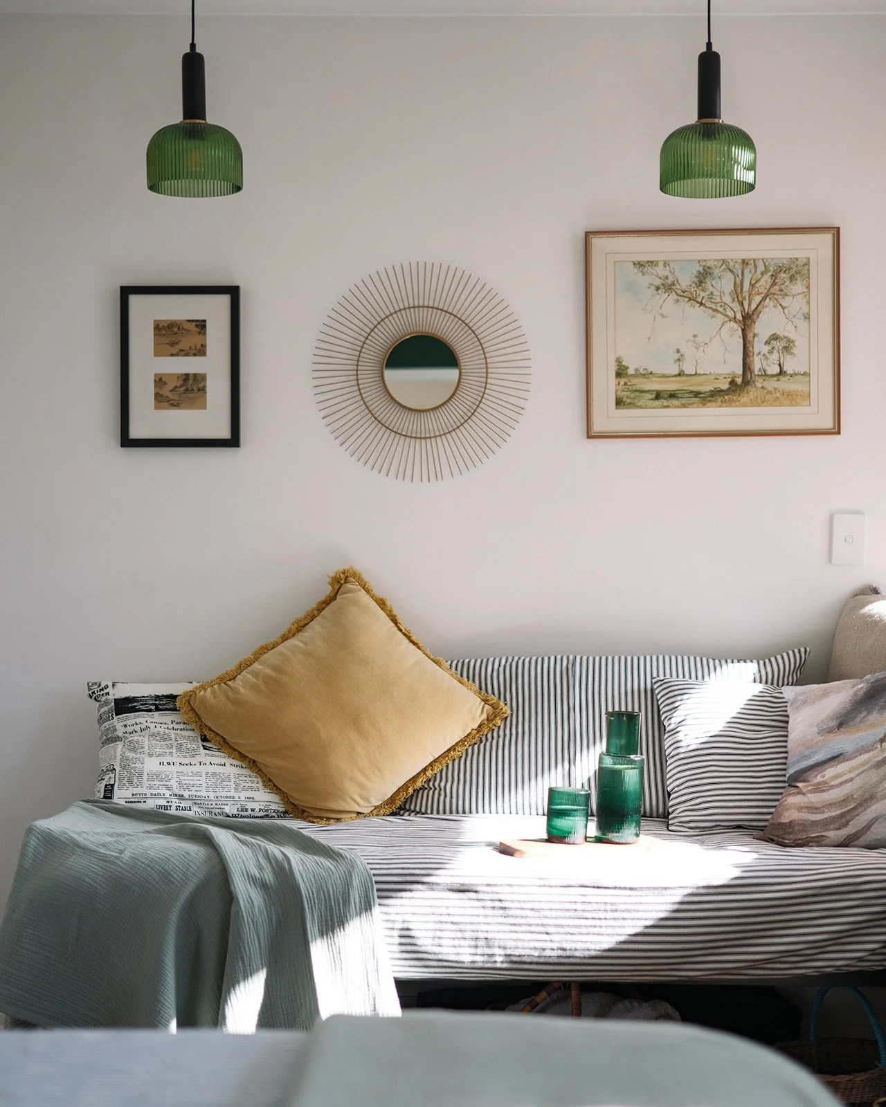 A cozy living room corner with a white wall decorated with picture frames, a round mirror, and landscape paintings. A beige pillow with tassels is on a striped sofa with black and white cushions. Green glass vases are on a small table, and sunlight c