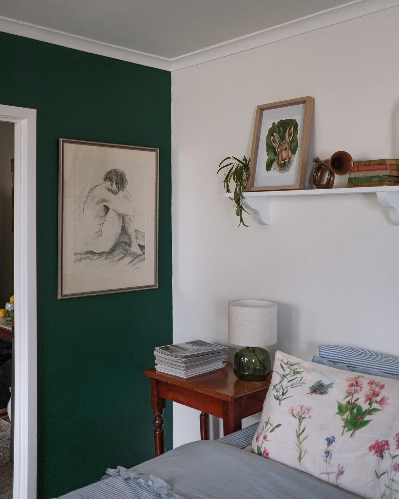 A cozy bedroom corner with a green accent wall, a framed sketch of a seated woman, a white shelf with a framed animal illustration, a potted plant, and decorative items, alongside a wooden nightstand with a stack of magazines and a glass lamp, and a 