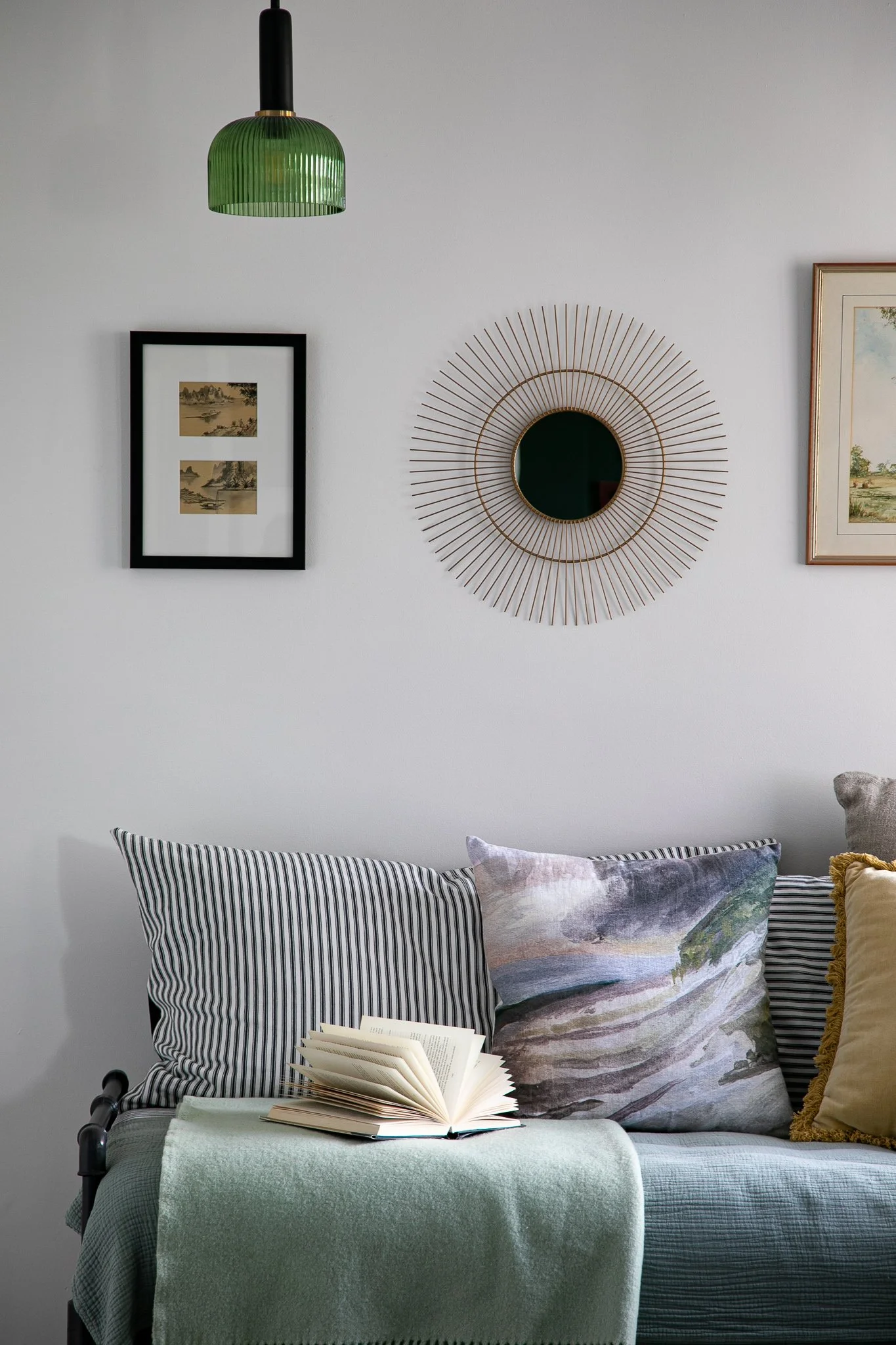 A living room with a white wall decorated with a round mirror with sunburst frame, framed landscape artwork, and a framed painting. Below, a gray sofa has striped, patterned, and plain cushions, with a light green throw blanket. On a side table, an o