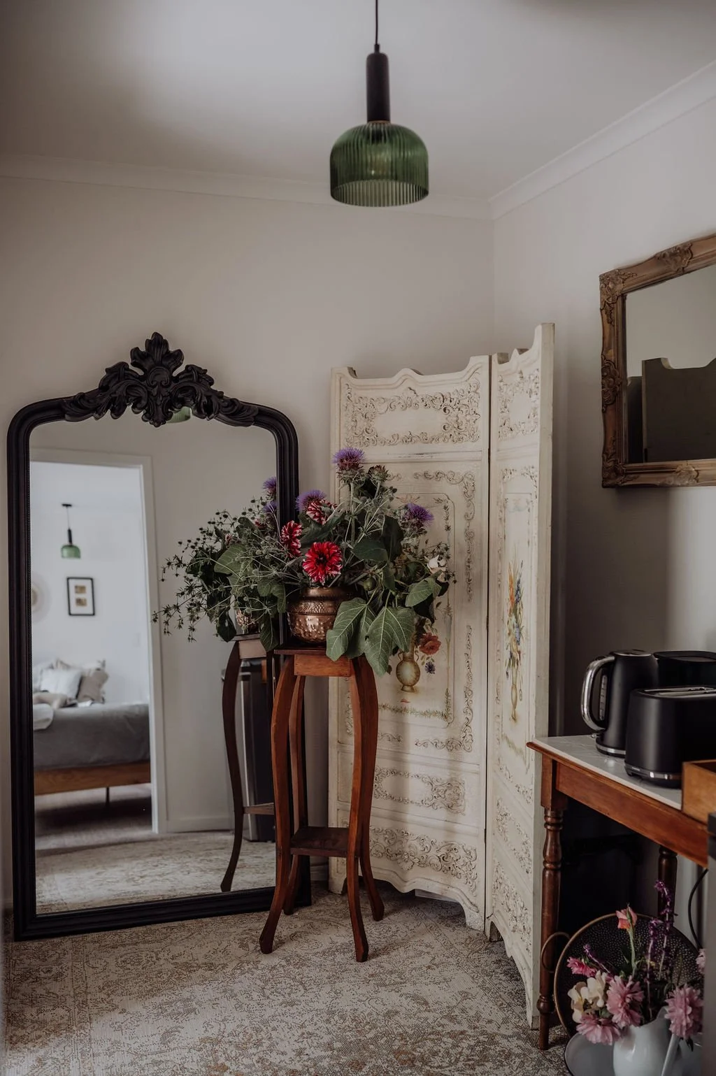 A cozy corner of a living space with a large, ornate mirror, floral arrangements, a white decorative folding screen, and a wooden table with black appliances.