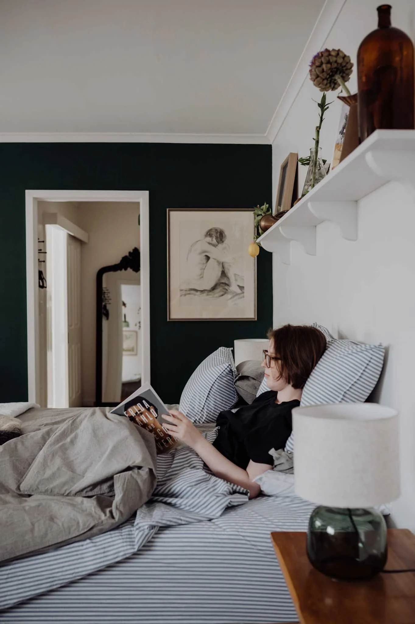 A woman with glasses and short hair lying in bed, reading a book, with striped bedding, a bedside table with a lamp, and a decorative glass container. Behind her, wall art and a shelf with a vase containing dried flowers are visible.