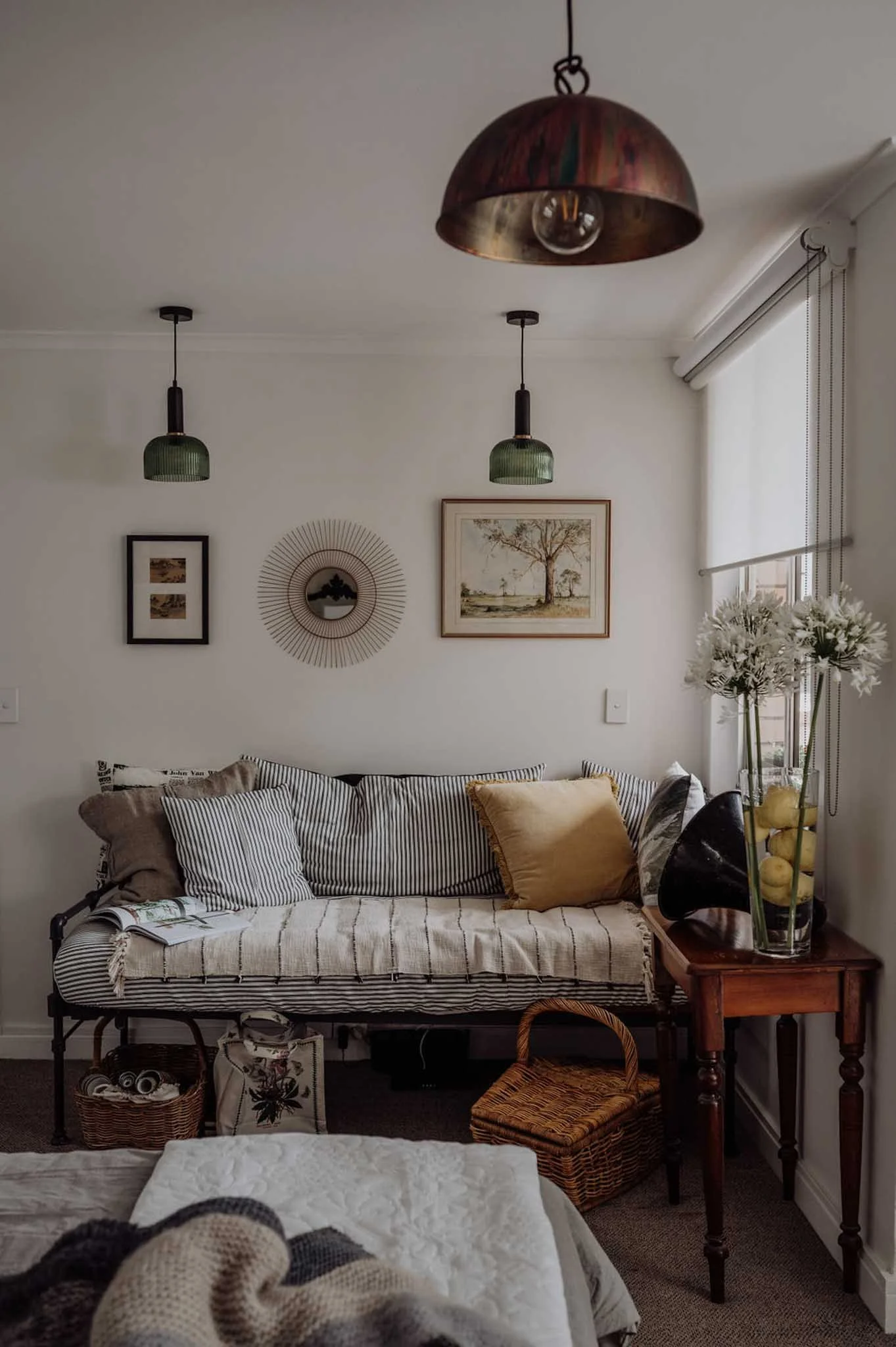 A cozy living room corner with a striped sofa adorned with pillows, framed artworks on the wall, pendant lights hanging from the ceiling, a side table with a vase of white flowers, and various baskets and decor items underneath.
