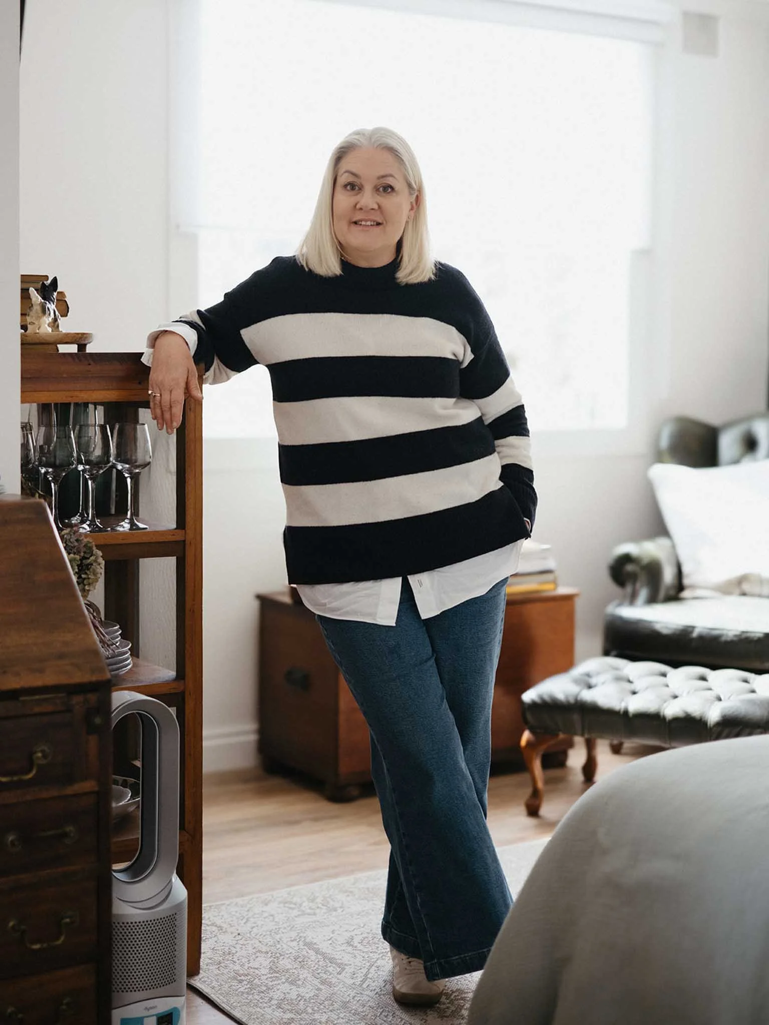 A woman with shoulder-length white hair, wearing a black and white striped sweater and blue jeans, standing in a living room with her hand resting on a wooden shelf, smiling at the camera.