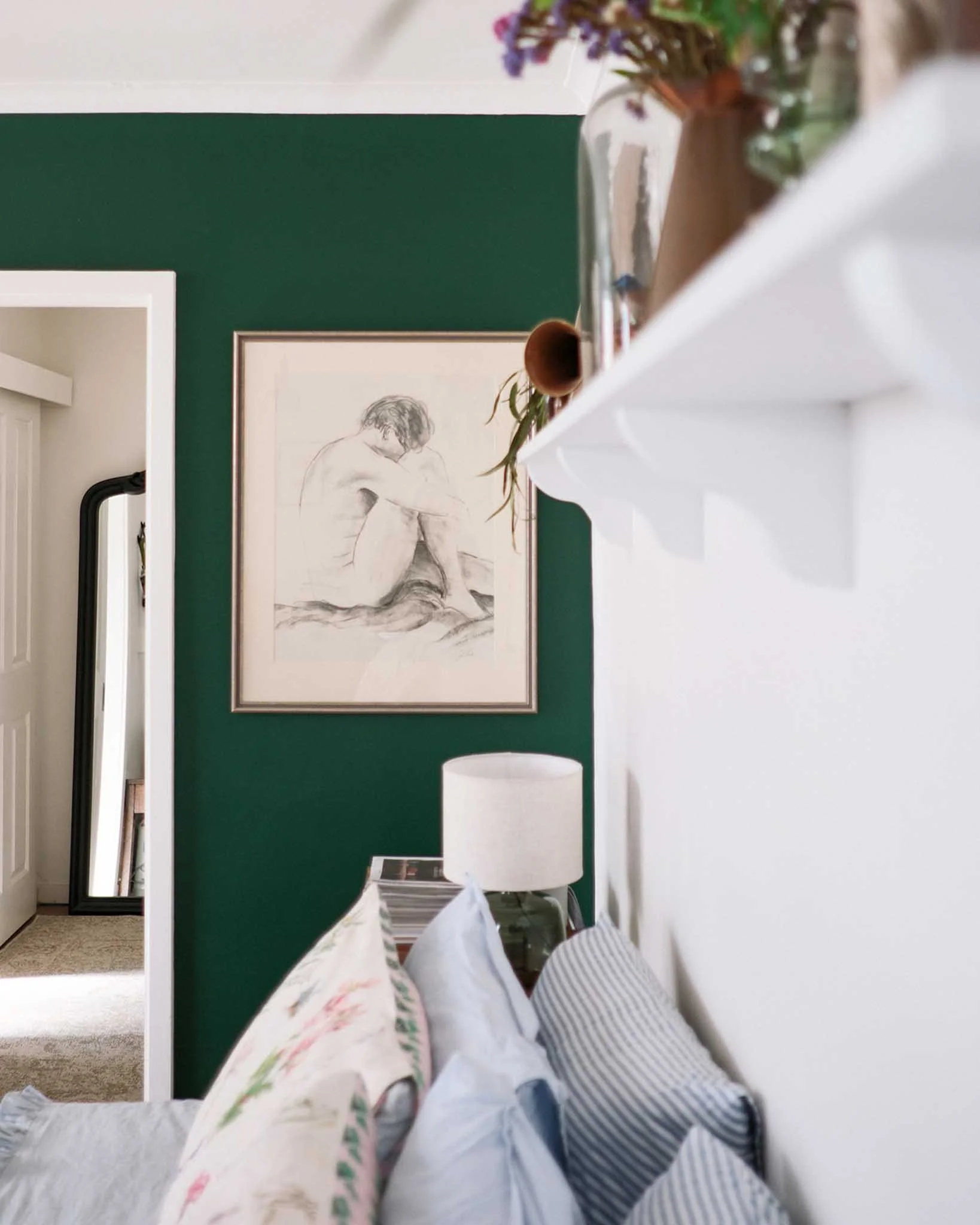 A room with a deep green wall featuring a framed sketch of a seated person with short hair. Partially visible white furniture and a tall white lamp are in the foreground. On the left, part of a mirror or window frame is visible, reflecting a portion 
