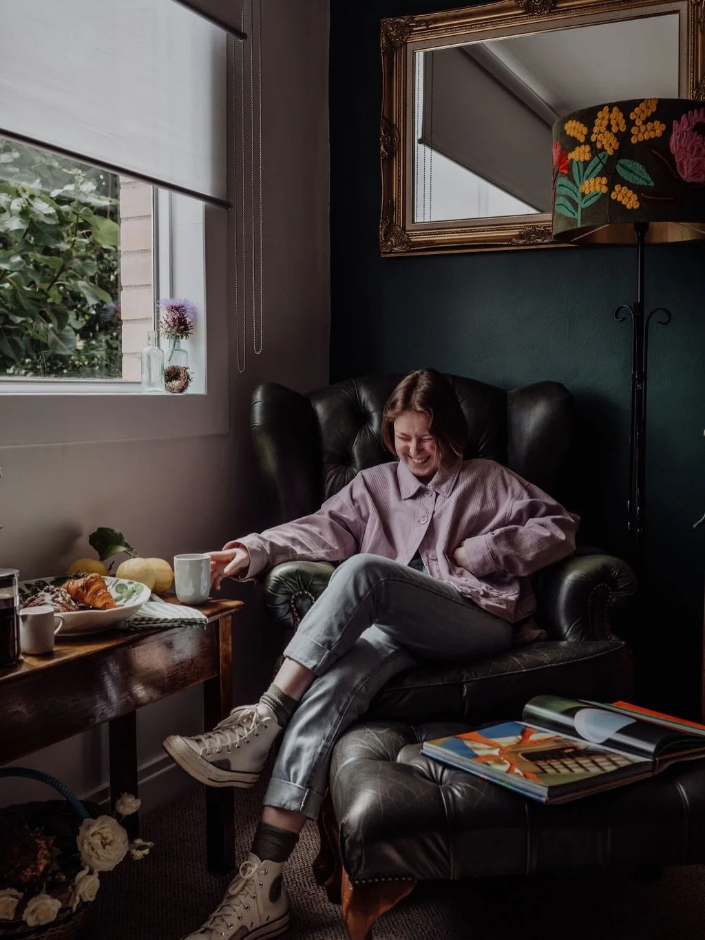 A young woman sitting in a black leather armchair, laughing and reaching for a white mug on a wooden table beside her. The table has a plate of croissants and a few pumpkins. A window beside her shows greenery outside, and there are decorative flower