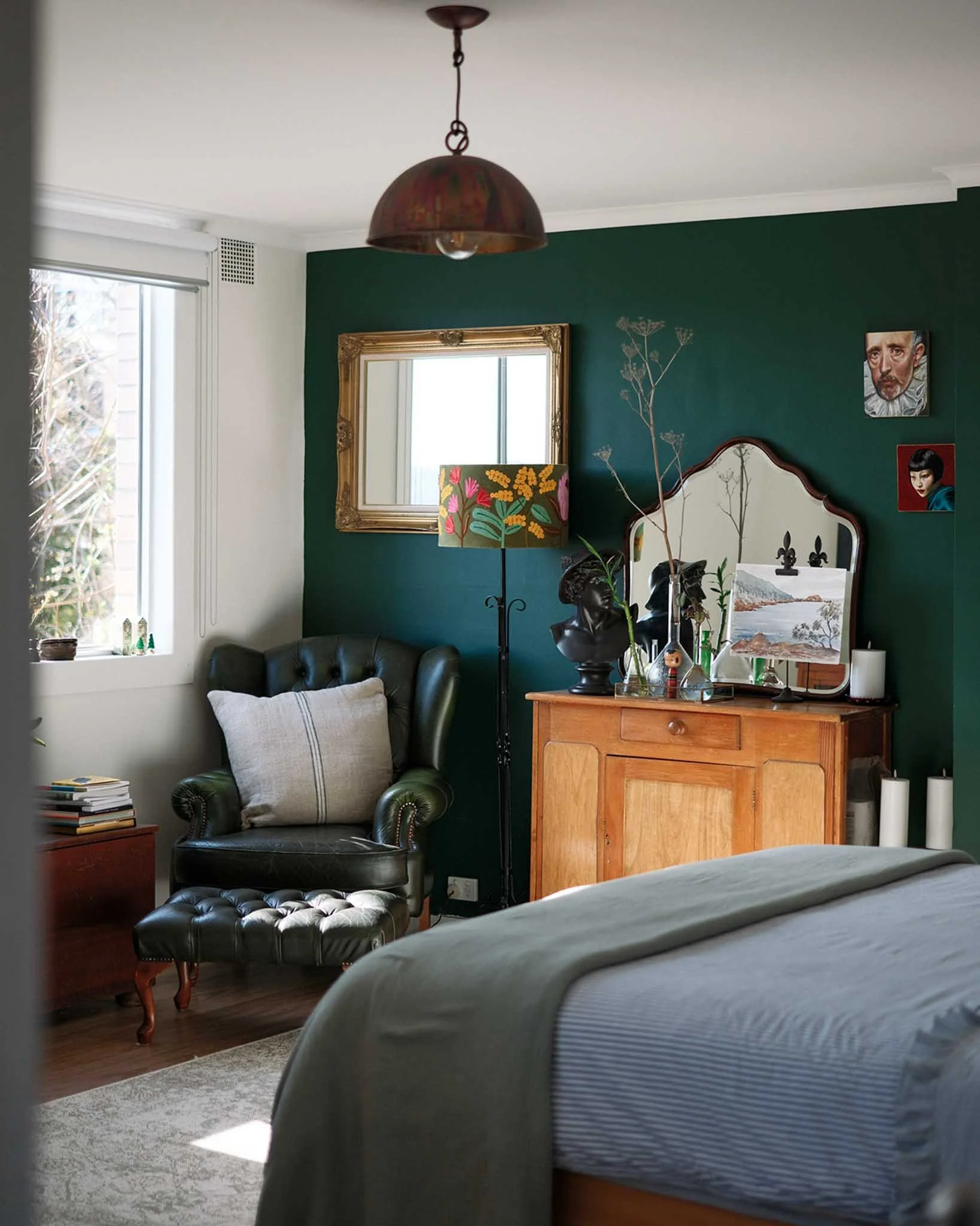 A cozy bedroom corner featuring a green accent wall, a gold framed mirror, a black leather armchair with a beige pillow, a wooden dresser with decorative items, and wall art including a portrait of a man and a woman, along with a window with blinds bringing in natural light.