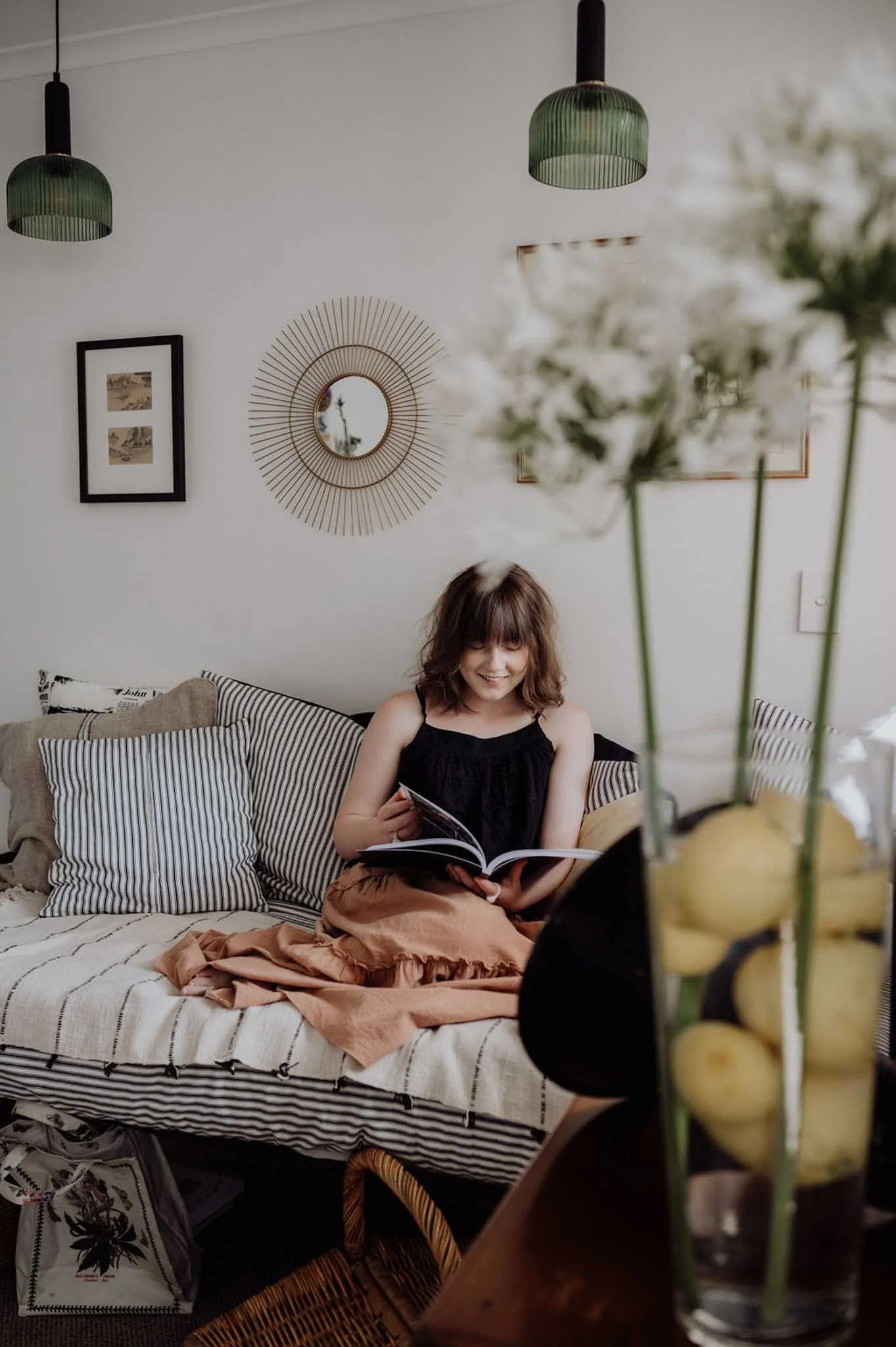 A woman with shoulder-length brown hair reading a book while sitting on a striped beige and white sofa in a cozy living room. The sofa has multiple striped and solid-colored pillows. Behind her, a white wall features framed art and a round decorative mirror. A glass vase with white flowers is in the foreground, slightly out of focus.