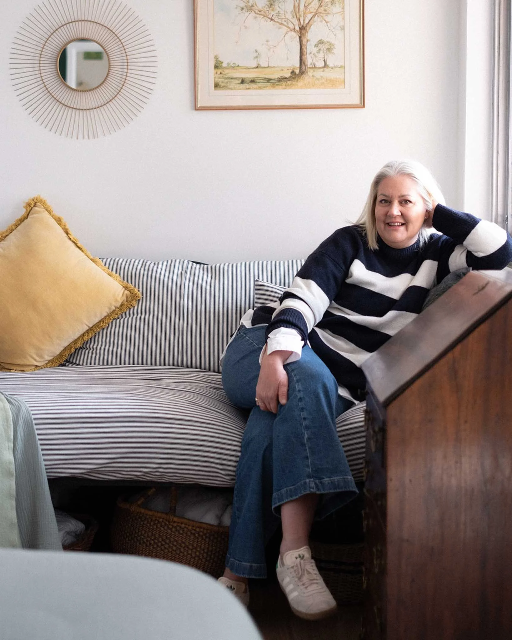 A woman with blonde hair sitting on a striped couch, smiling, wearing a black and white striped sweater, blue jeans, and white sneakers. There is a large yellow pillow on the couch, and behind her are picture frames and a window with a curtain. The wall has a decorative round mirror and a framed painting of trees.