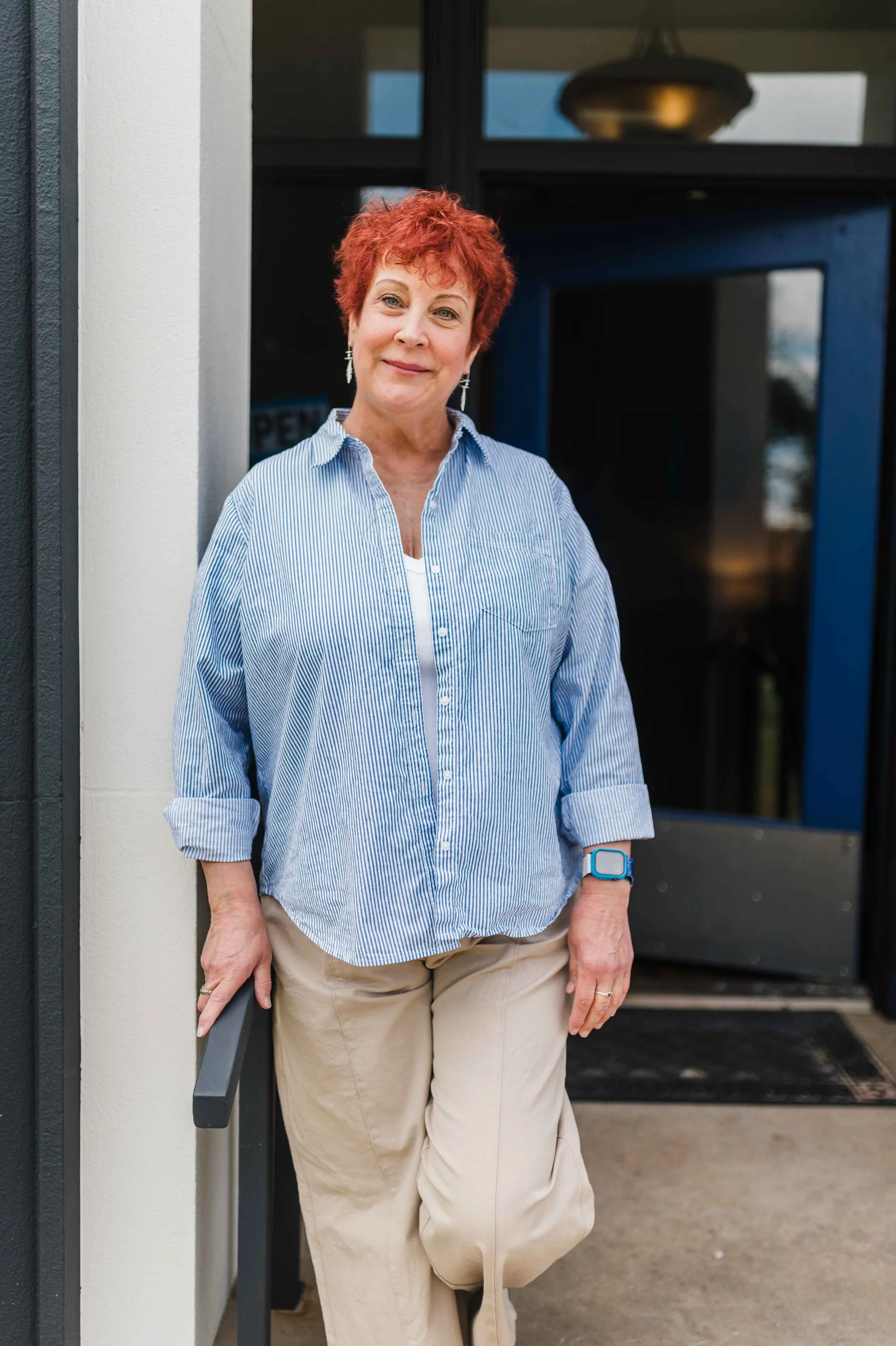 A woman with short red curly hair, wearing a blue and white striped shirt, beige pants, and a smartwatch on her left wrist, standing outside a building with a dark doorway.