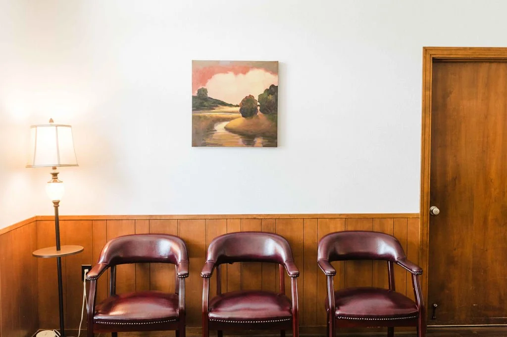 Three maroon chairs in front of a wooden-paneled wall with a landscape painting above, a wooden door to the right, and a tall standing lamp on the left.