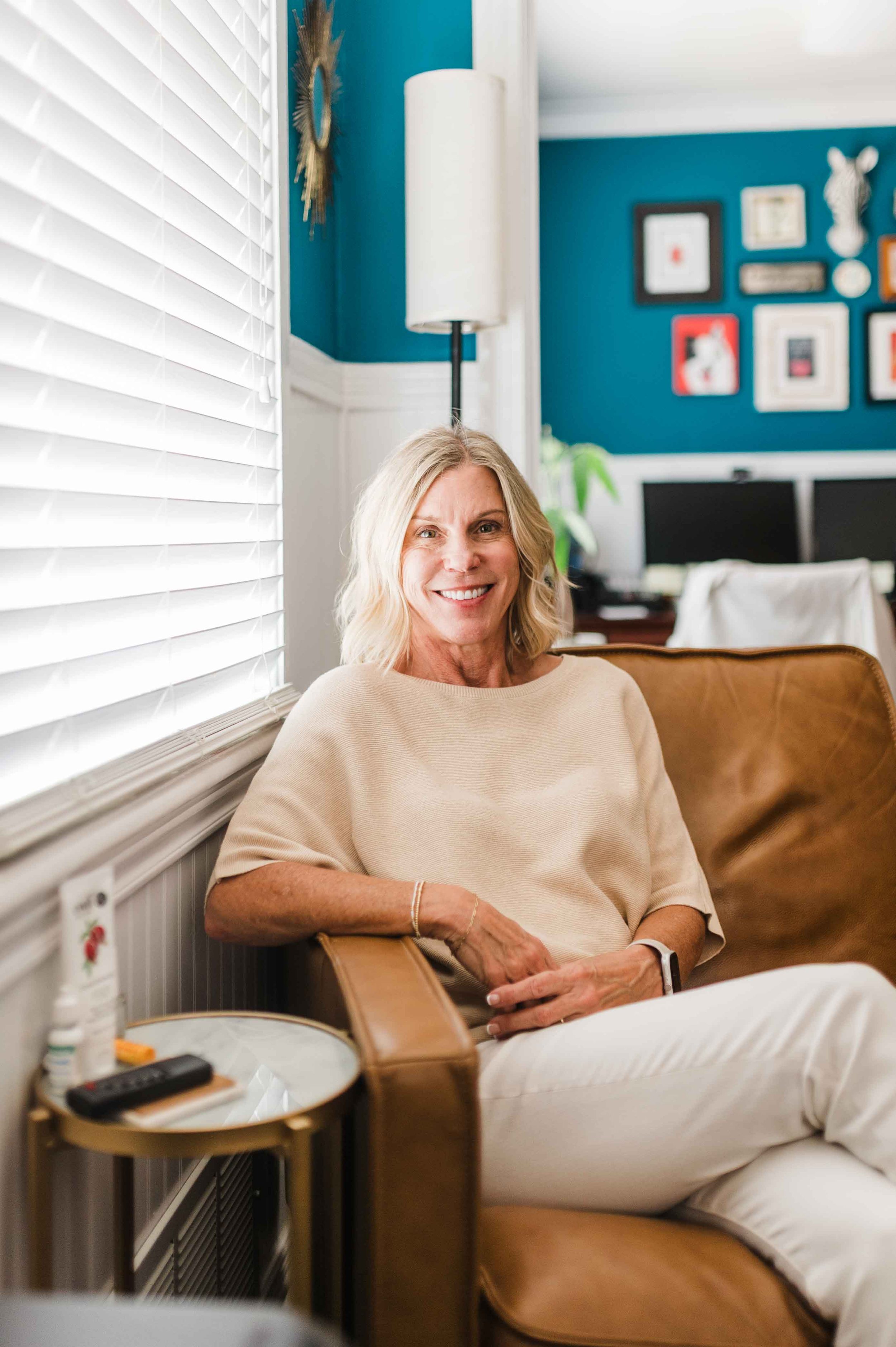 A woman with blonde hair and a beige shirt sitting on a couch in a room with teal walls, a gallery wall of framed artwork, and a white lamp.