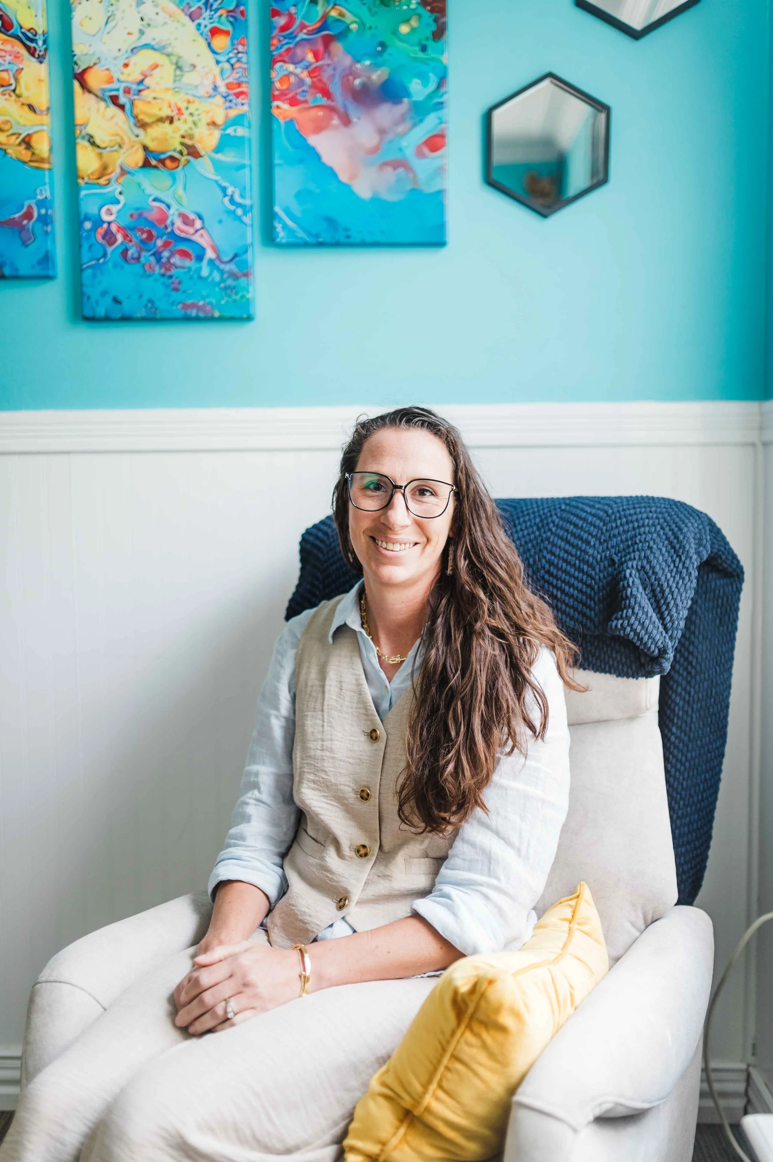 A woman with glasses and long braided hair, smiling, sitting on a white armchair in a room with blue walls, colorful abstract paintings, and hexagonal mirrors.