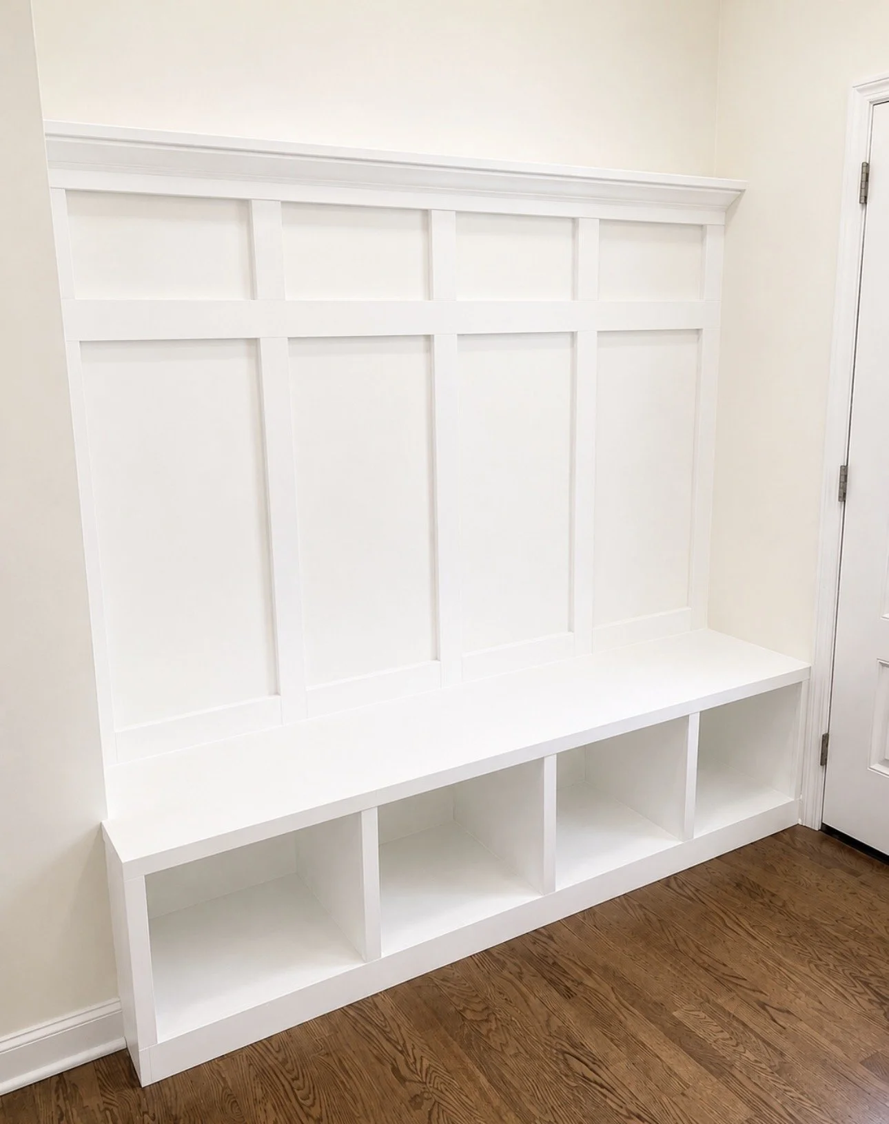 White built-in bench with a cubby storage unit and decorative paneling on the back, next to a door with a silver hinge on a wood floor.