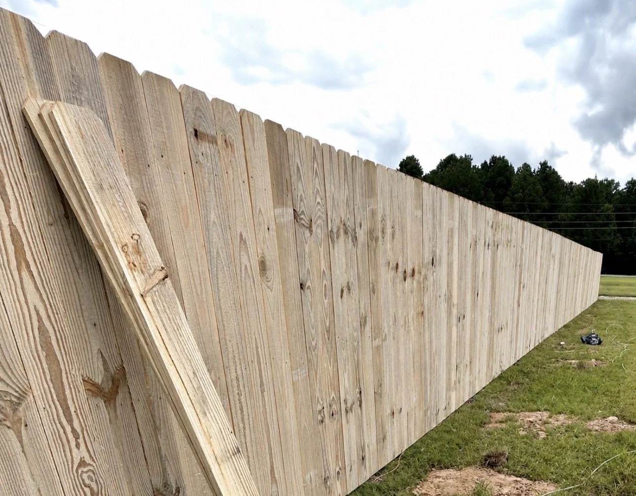 Long wooden fence under construction on grassy area with trees and cloudy sky in the background.