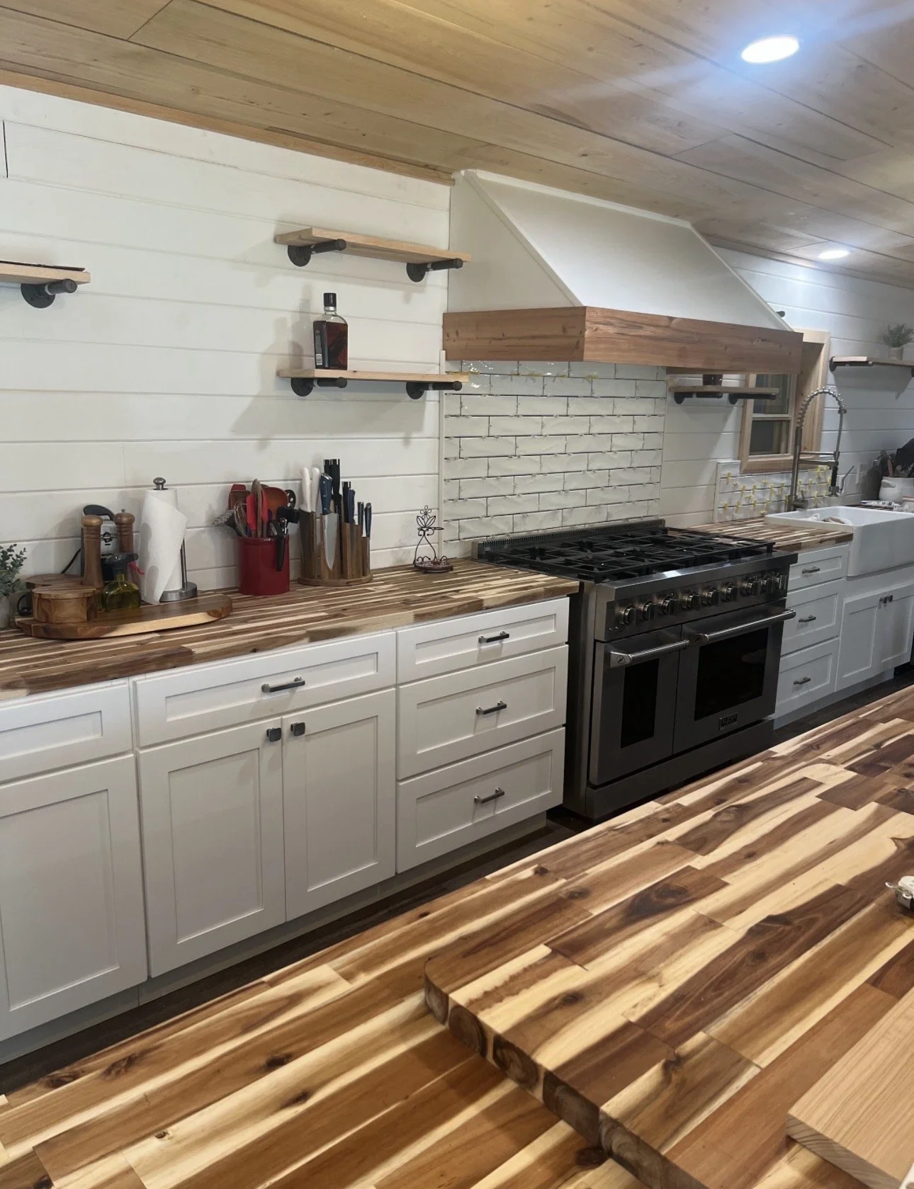 A modern kitchen with wooden countertops, white cabinets, and a black stove. Open shelving with small decor and utensils, a white brick backsplash, and a farmhouse sink with a kitchen faucet. Wooden flooring and ceiling with recessed lighting.