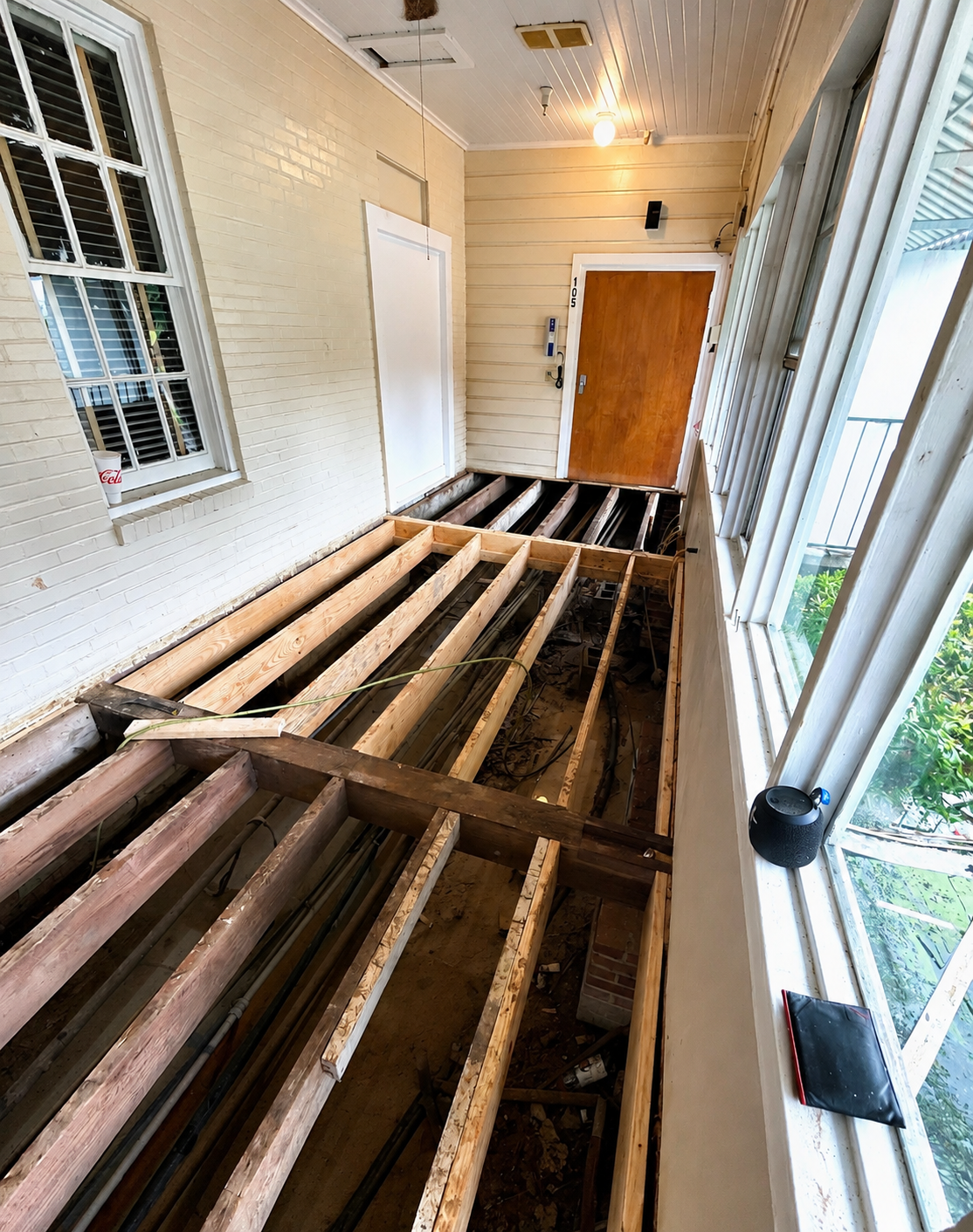 Porch with flooring under renovation, showing exposed wooden floor joists, a white brick wall with window, and a wooden door at the end of the porch.