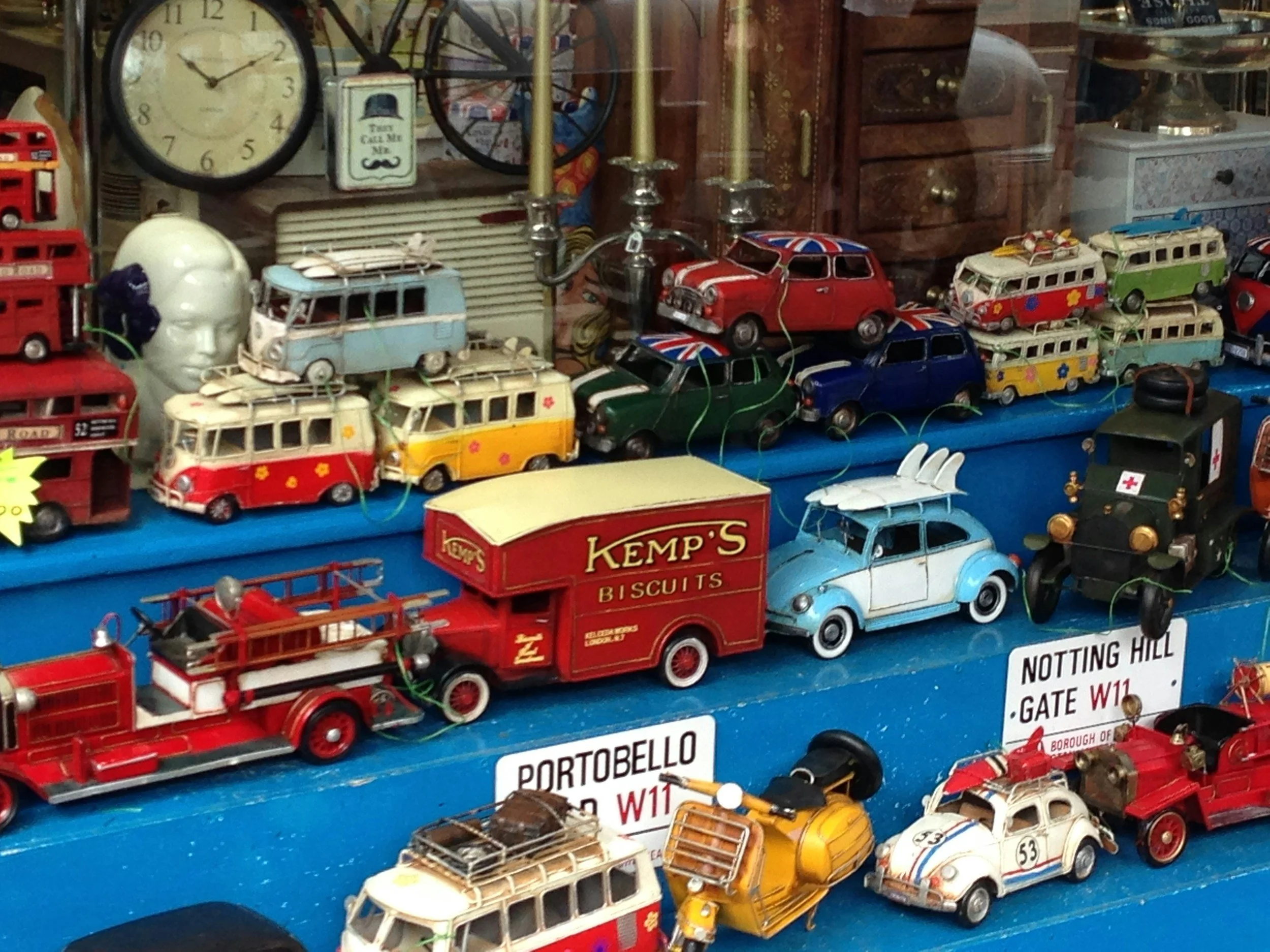 A display of vintage toy cars and trucks, including miniature vehicles resembling buses, a fire truck, a car with Union Jack flags, and other classic car models, arranged on a blue surface with aisle signs in the background.