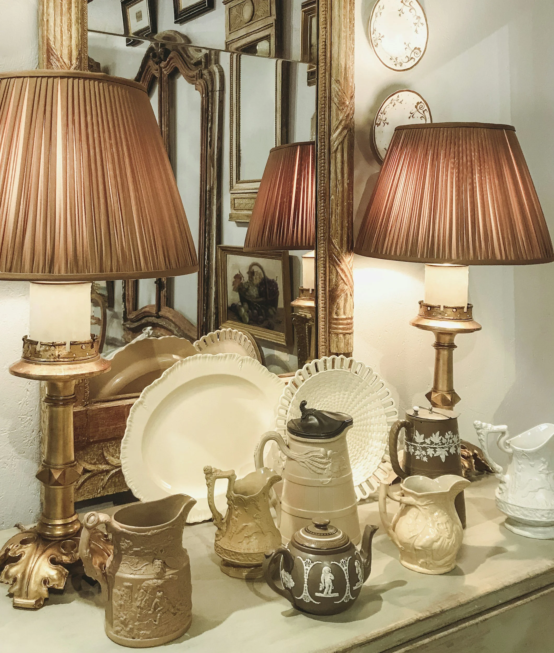 Display of various vintage and antique ceramic and porcelain vases, pitchers, and decorative dishes, arranged on a surface with a large ornate mirror and framed artwork in the background.