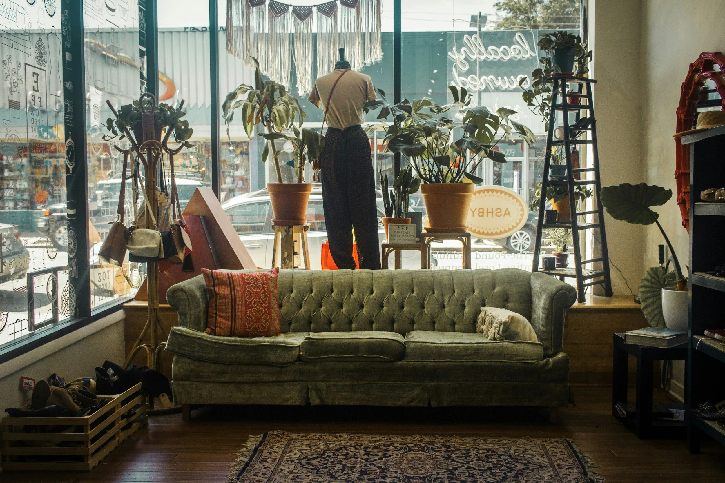 The image depicts the interior of a cozy shop with a vintage style. In the foreground, there is a green, tufted sofa with patterned cushions. Behind the sofa, a large window allows natural light to fill the space, showcasing various indoor plants, including a large potted plant and smaller plants. There is a mannequin dressed in a casual outfit and some decorative furniture, like a small table with a sign reading ‘Please ask for assistance’ and a vintage rug on the wooden floor. Other store items, such as hats, shoes, and shelving units, are visible around the space.