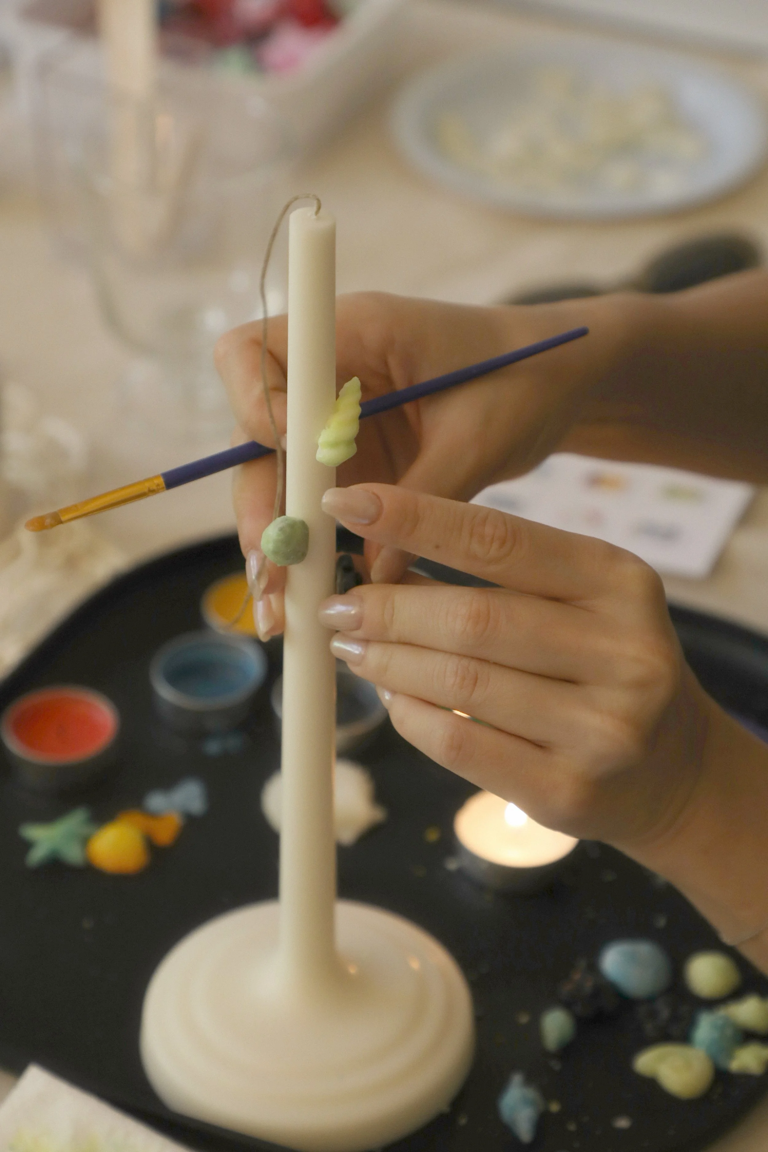 Close-up of a person's hand holding a long white candle with small dollops of green wax on it, and a thin blue stick with a small amount of yellow wax. In the background, there is a black board with watercolor paints and some scattered small colorful wax blobs.