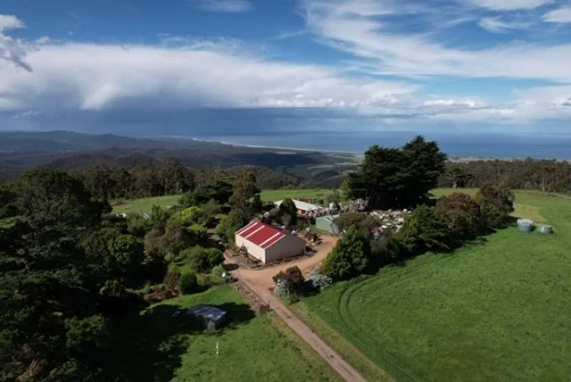A rural landscape featuring a farm with several buildings, surrounded by green fields, trees, and a distant view of the ocean under a partly cloudy sky.