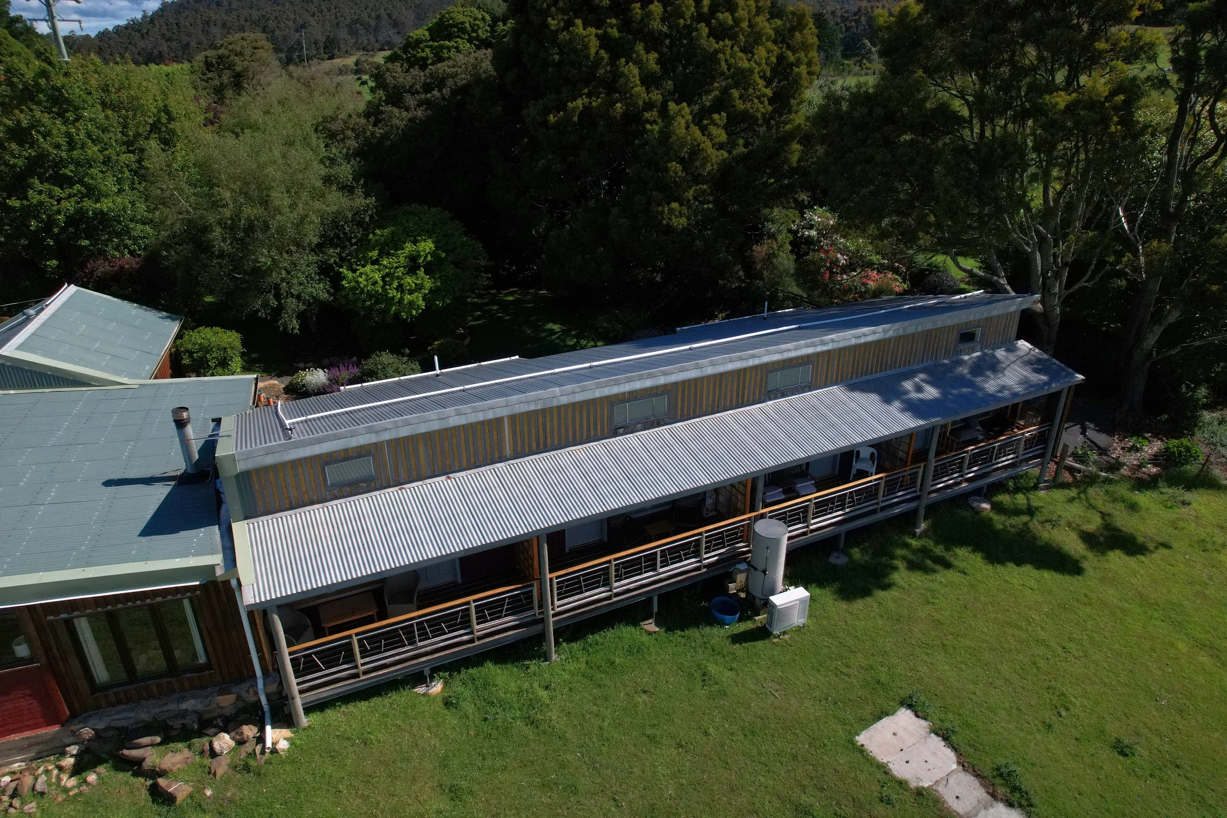 An aerial view of a modern building with metal roofing and large windows, surrounded by lush green trees and a manicured lawn in a hilly area.