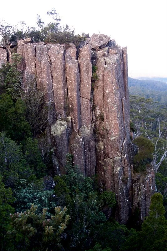 Large vertical rock formation with a flat top, surrounded by dense green trees and shrubs.
