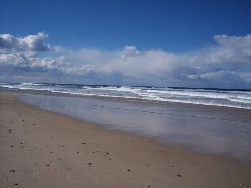 Empty beach with wet sand, gentle waves, and blue sky with scattered clouds.