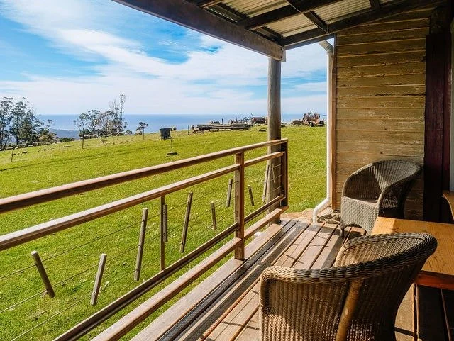 A view from a covered porch overlooking a grassy field with trees, pastures, and the ocean in the distance.
