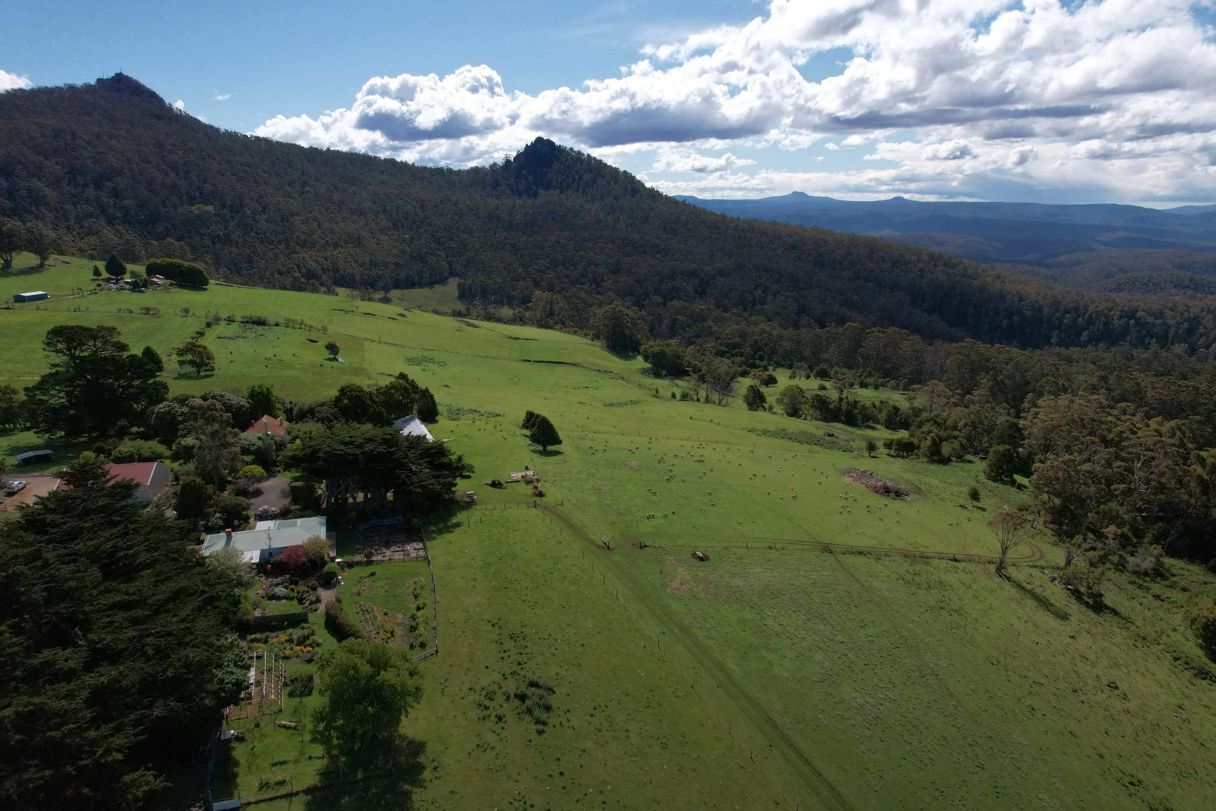 A scenic landscape showing green rolling hills, a few houses and trees, with a mountain range in the background under a partly cloudy sky.