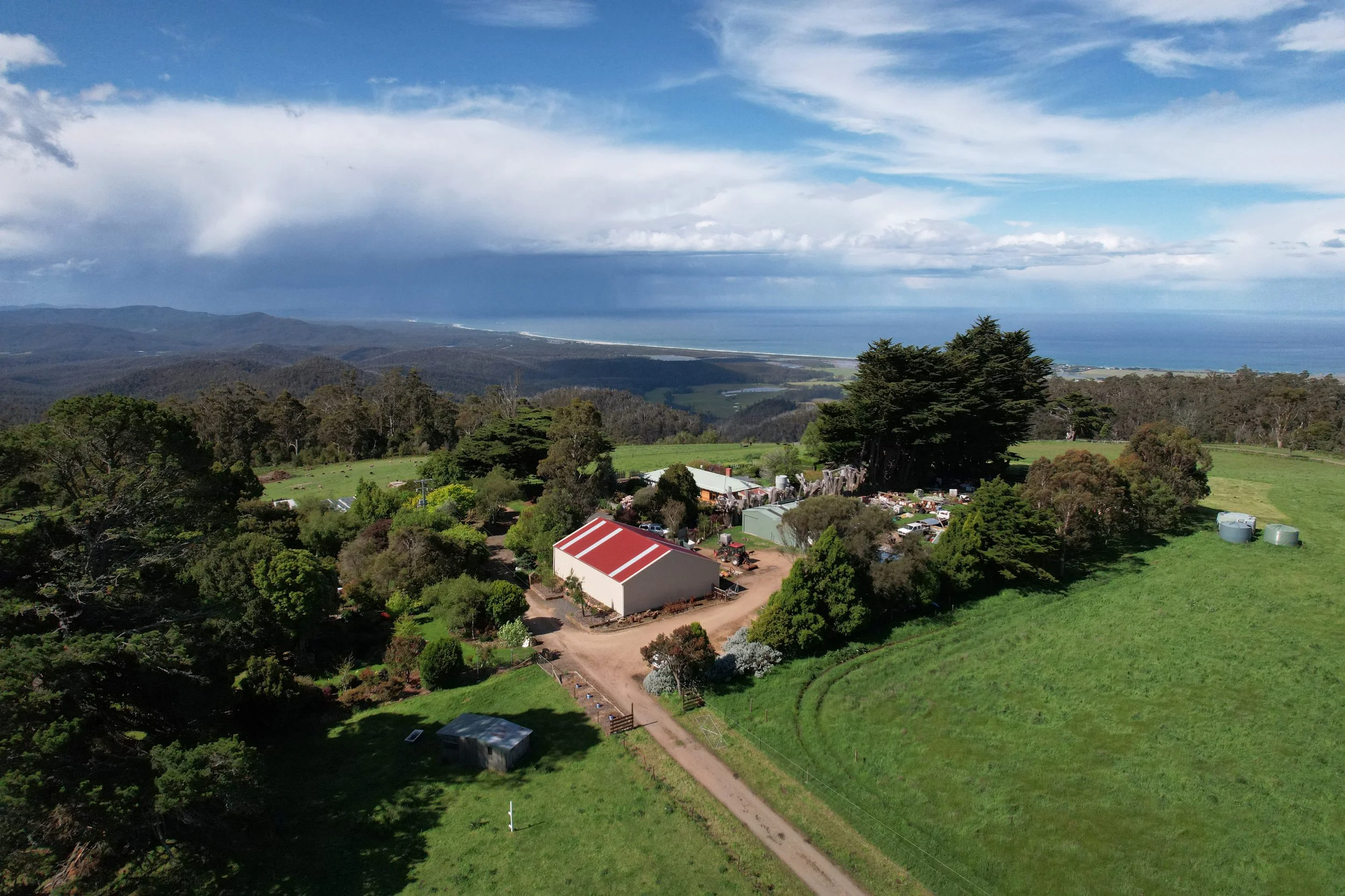 Aerial view of a rural landscape with lush green fields, trees, a red-roofed building, and a winding dirt road, overlooking distant mountains and a cloudy sky over the ocean.
