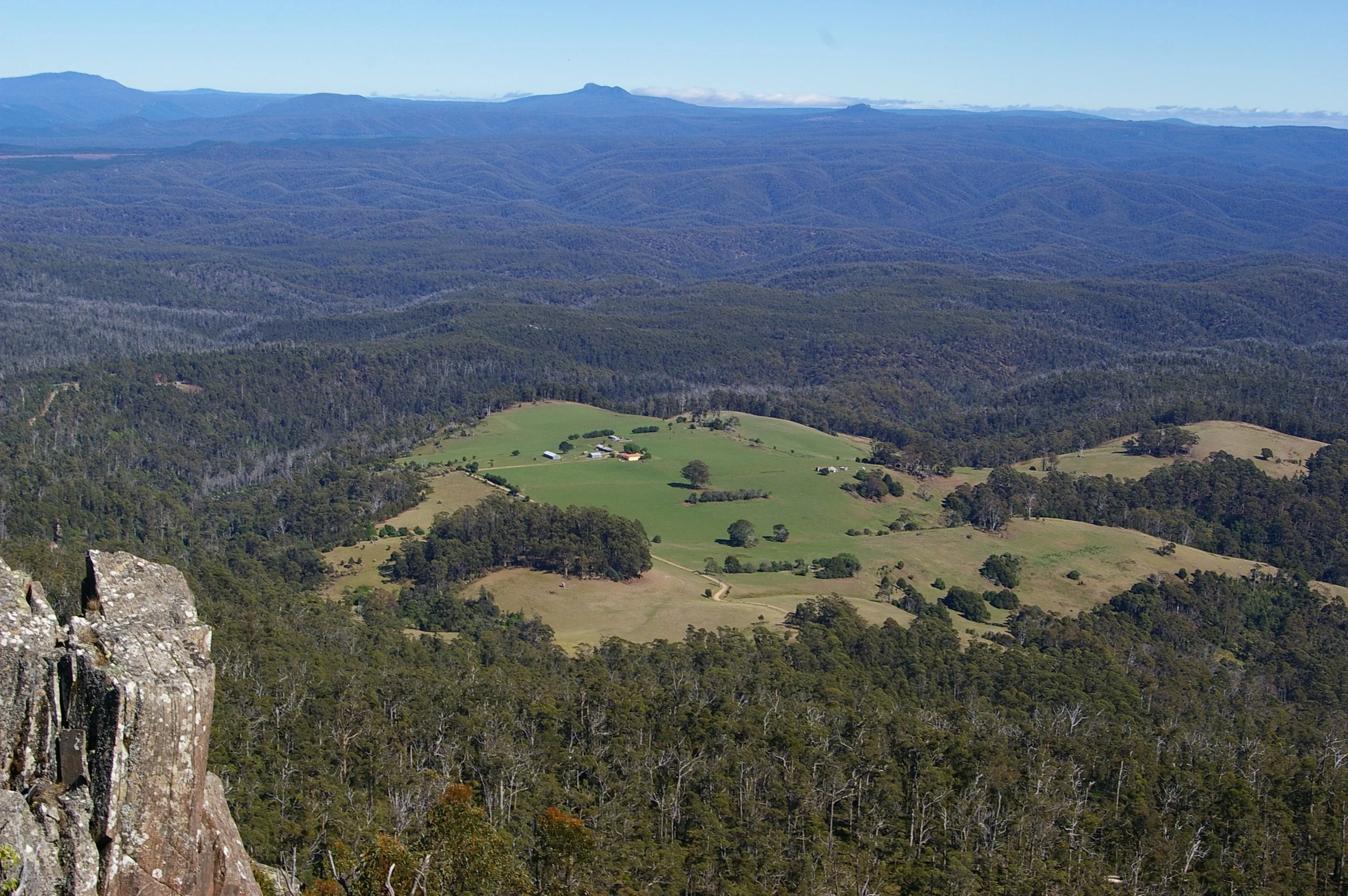 Mountain landscape with forested hills and a green valley with farm buildings, under a clear blue sky.