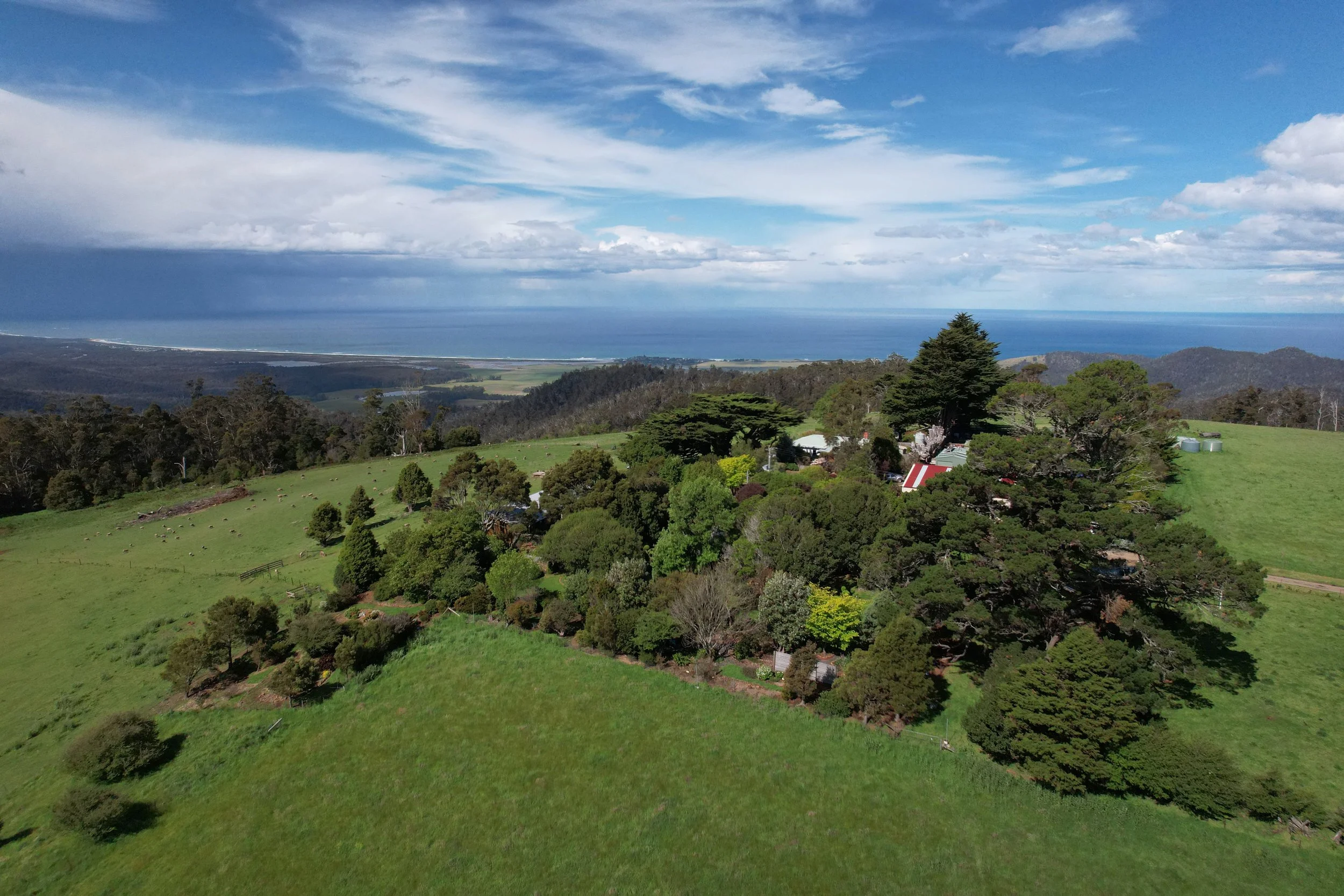 Scenic view of rolling green hills and lush trees with a few buildings, overlooking a distant ocean under partly cloudy sky.