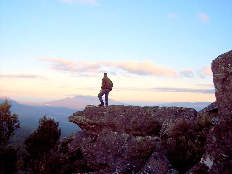 A person standing on a large rock formation overlooking a valley with mountains in the distance during sunset.
