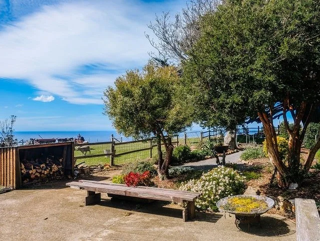 Outdoor garden area with wooden benches, trees, flowers, and a view of the ocean in the background.