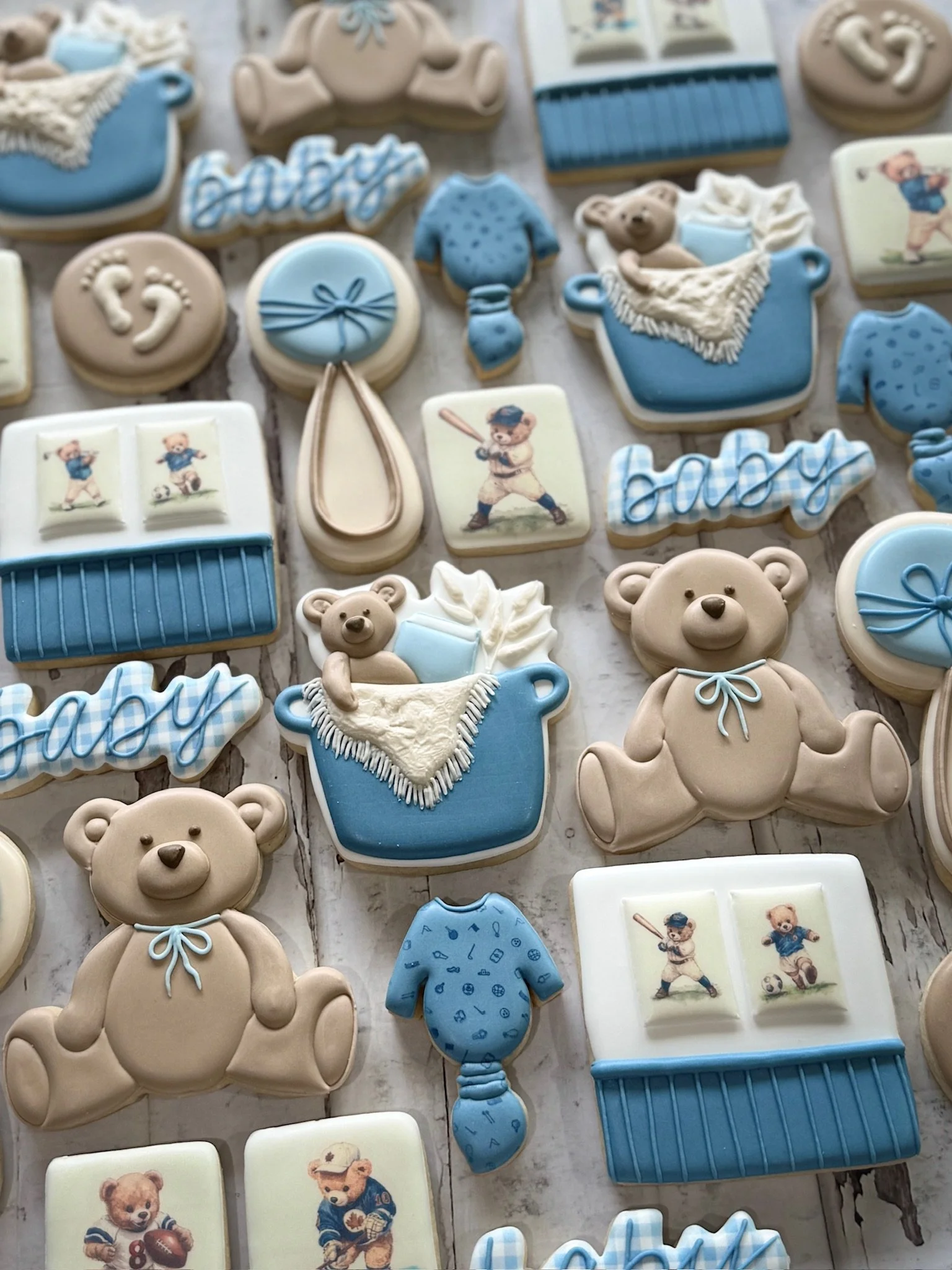 Assortment of blue and beige baby shower cookies decorated with teddy bears, onesies, rattles, baby bottles, footprints, and the name “Blaise.”