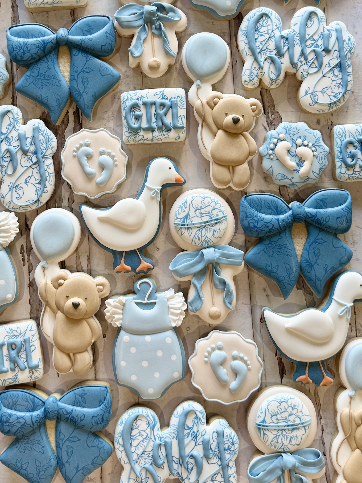 Blue and white baby-themed cookies, including bows, teddy bears, ducks, onesies, pacifiers, footprints, and floral name plaques.